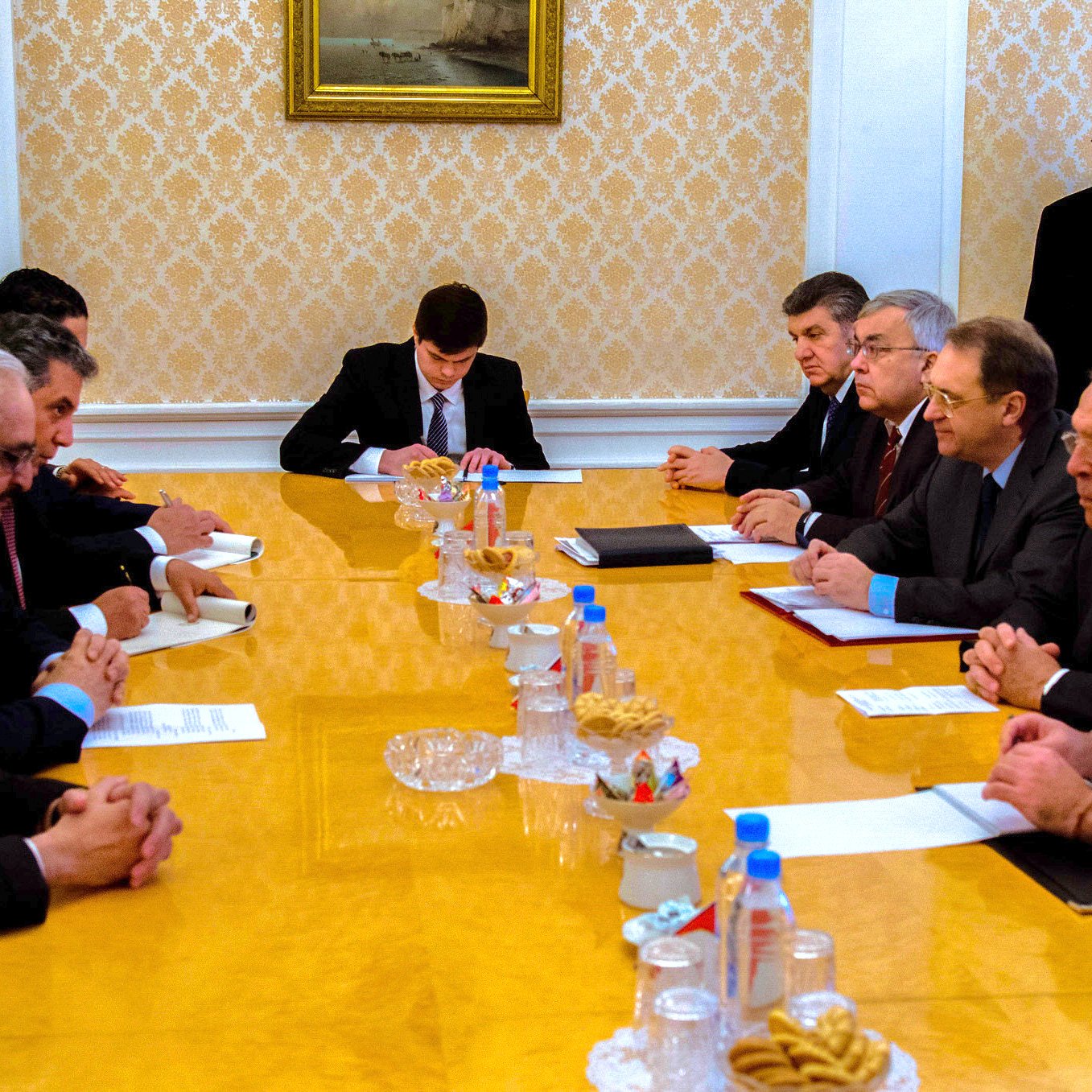 The image depicts a formal meeting taking place around a long wooden table in what appears to be a conference room. Participants are seated on both sides of the table, engaged in conversation or listening attentively. The table is set with various documents, and there are snacks and beverages in front of some attendees. The room has an elegant decor with a patterned wall and paintings. A few individuals are standing in the background, likely observing the meeting. The atmosphere seems serious and focused on discussion.