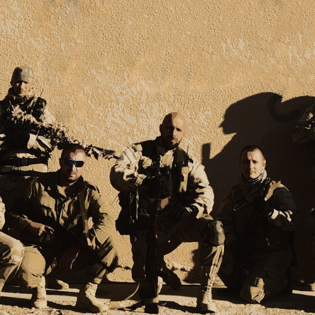 L'immagine mostra un gruppo di otto soldati in divisa militare, affiancati e in posa. Sono armati e sembrano essere in un ambiente desertico o in una zona di conflitto. I soldati indossano attrezzature tattiche e occhiali da sole, e la luce evidenzia i dettagli delle loro uniformi. Sullo sfondo si vede una parete di un edificio, suggerendo un contesto operativo. L'atmosfera è seria e professionale.