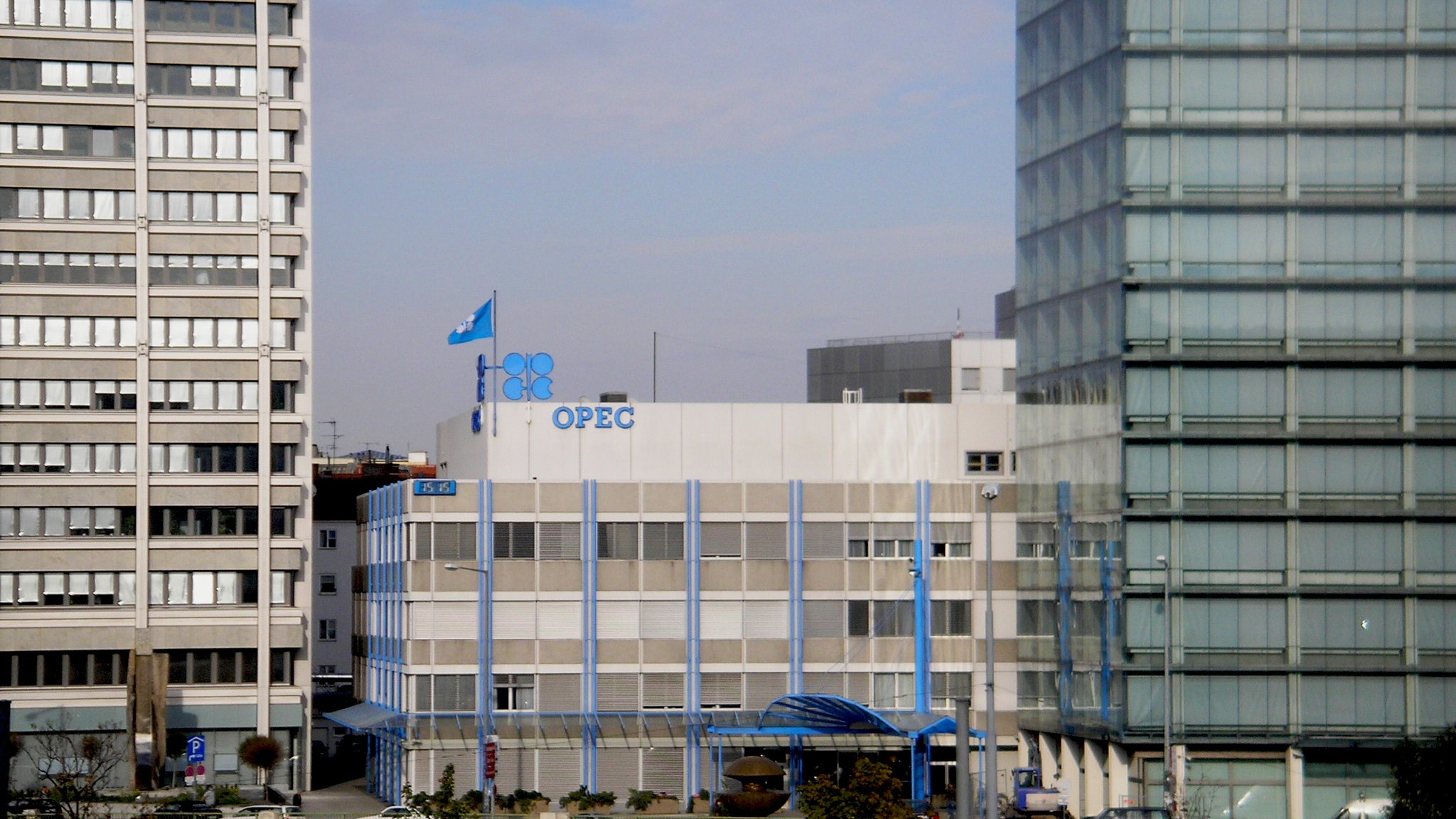 L'image montre le bâtiment de l'OPEP (Organisation des pays exportateurs de pétrole), qui est un bâtiment moderne à plusieurs étages. Des drapeaux sont visibles sur le toit, y compris celui de l'OPEP. Le bâtiment est entouré par d'autres structures urbaines, reflétant un environnement de ville. Le ciel est légèrement nuageux, suggérant une journée calme. Des éléments architecturaux en verre et en béton contrastent avec le design du bâtiment de l'OPEP.