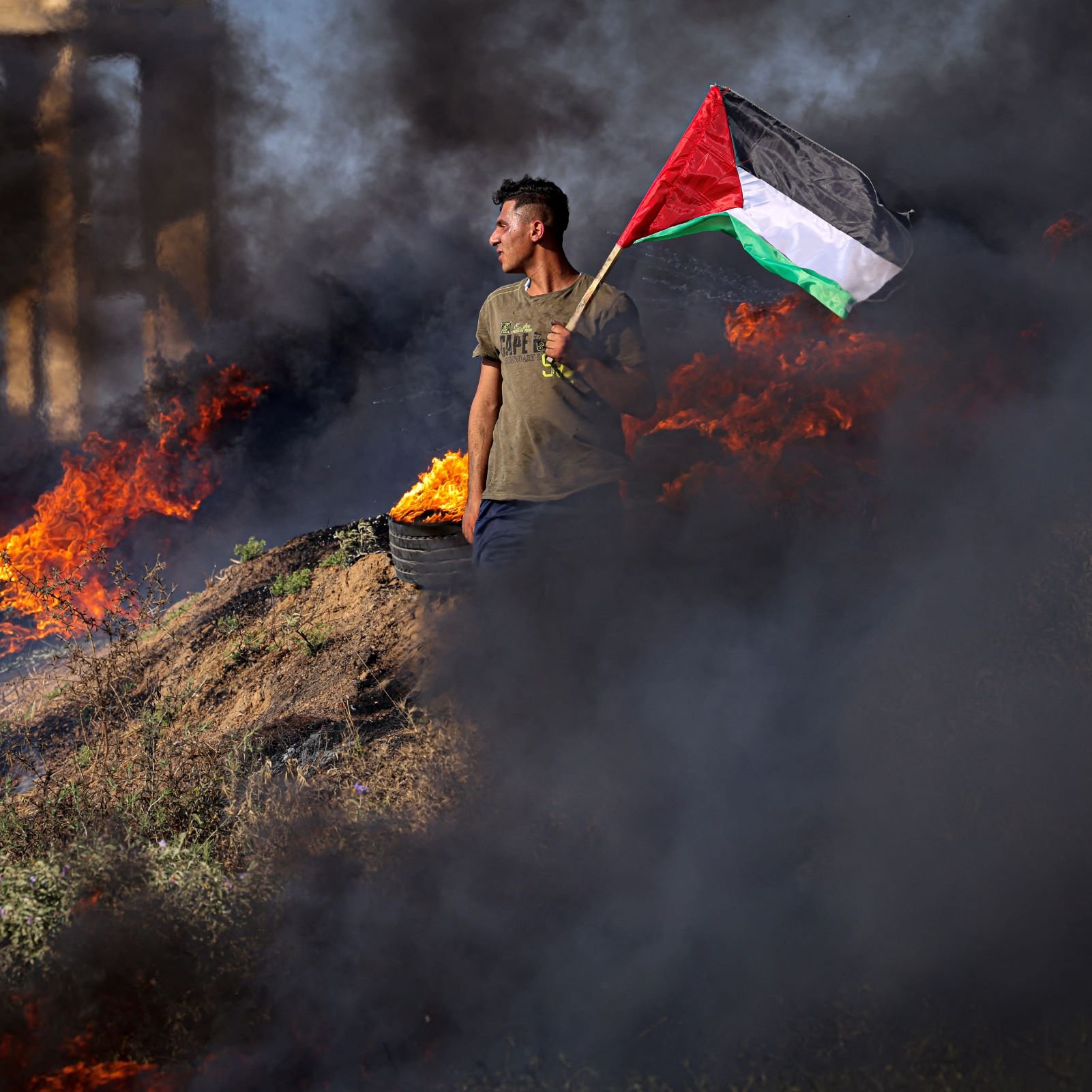 L'image montre une personne debout sur un terrain où des feux brûlent, produisant une épaisse fumée noire. Cette personne tient fièrement un drapeau palestinien, qui est composé de bandes rouge, noire, blanche et verte. Le paysage semble chaotique en raison des flammes entourant le manifestant, créant une atmosphère intense. Les éléments visuels de l'image évoquent des sentiments de militantisme et de détermination.