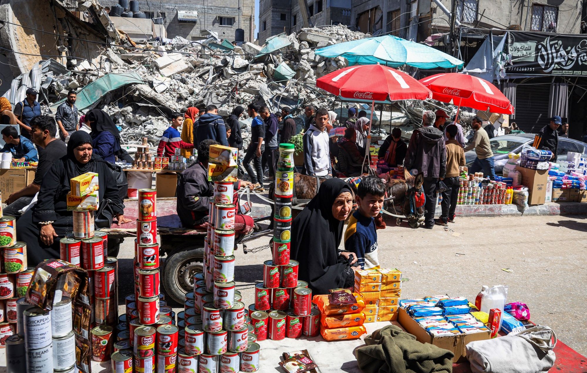 L'image montre une scène d'un marché en plein air, où des gens se déplacent parmi des étals de produits. Au premier plan, une femme en hijab et un enfant sont assis devant une pile de conserves et d'autres produits alimentaires. En arrière-plan, des bâtiments en ruines et des débris sont visibles, suggérant un contexte de destruction. Des parasols de différentes couleurs offrent de l'ombre aux vendeurs et aux clients. L'atmosphère semble chargée de vie malgré le décor dévasté.