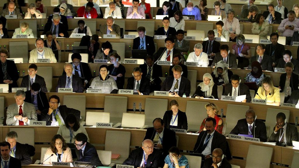 L'image montre une grande salle de conférence remplie de personnes assises à des tables. Les participants semblent concentrés sur leurs ordinateurs portables et divers documents, suggérant qu'ils sont engagés dans une discussion ou une réunion importante. À l'arrière-plan, on peut voir des drapeaux de différents pays visibles, ce qui indique un contexte international. L'éclairage de la salle est tamisé, créant une atmosphère sérieuse et studieuse.