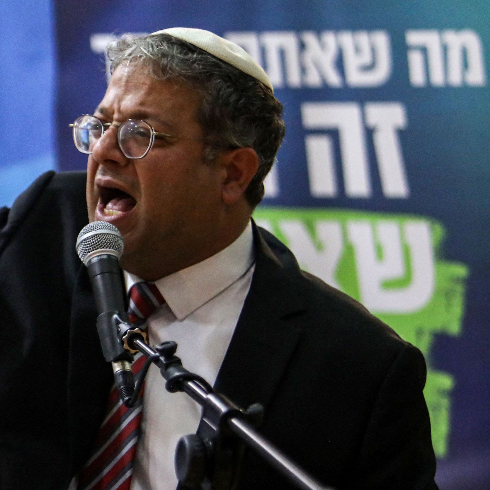 در این تصویر، یک مرد با عینک و کلاهی روی سر در حال صحبت کردن است. او به میکروفون نزدیک شده و به نظر می‌رسد که passionately در حال بیان نقطه نظری است. در پس‌زمینه، تعدادی لوگو و نوشته‌های مربوط به رسانه‌ها دیده می‌شوند. به نظر می‌رسد این نشست یا سخنرانی از اهمیت خاصی برخوردار باشد.