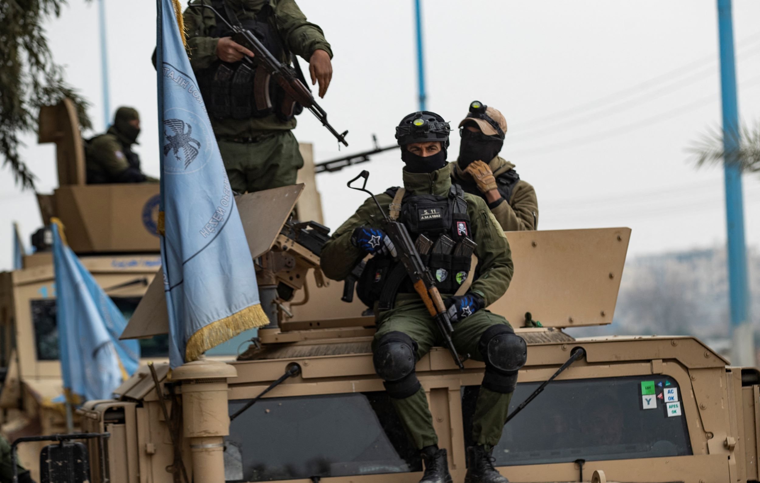 تظهر في الصورة مجموعة من الجنود العسكريين أثناء وجودهم في مركبة مدرعة. يرتدي الجنود زيًا عسكريًا ويظهر على بعضهم معدات تكتيكية وأسلحة. أحد الجنود يجلس على الجزء العلوي من المركبة، بينما يظهر آخرون يقفون عليها. هناك علم أزرق يعود لجهة معينة يبدو أنه متعلق بمهام حفظ السلام. الأجواء المحيطة تبدو حضرية مع وجود أشجار ومرافق عامة في الخلفية.