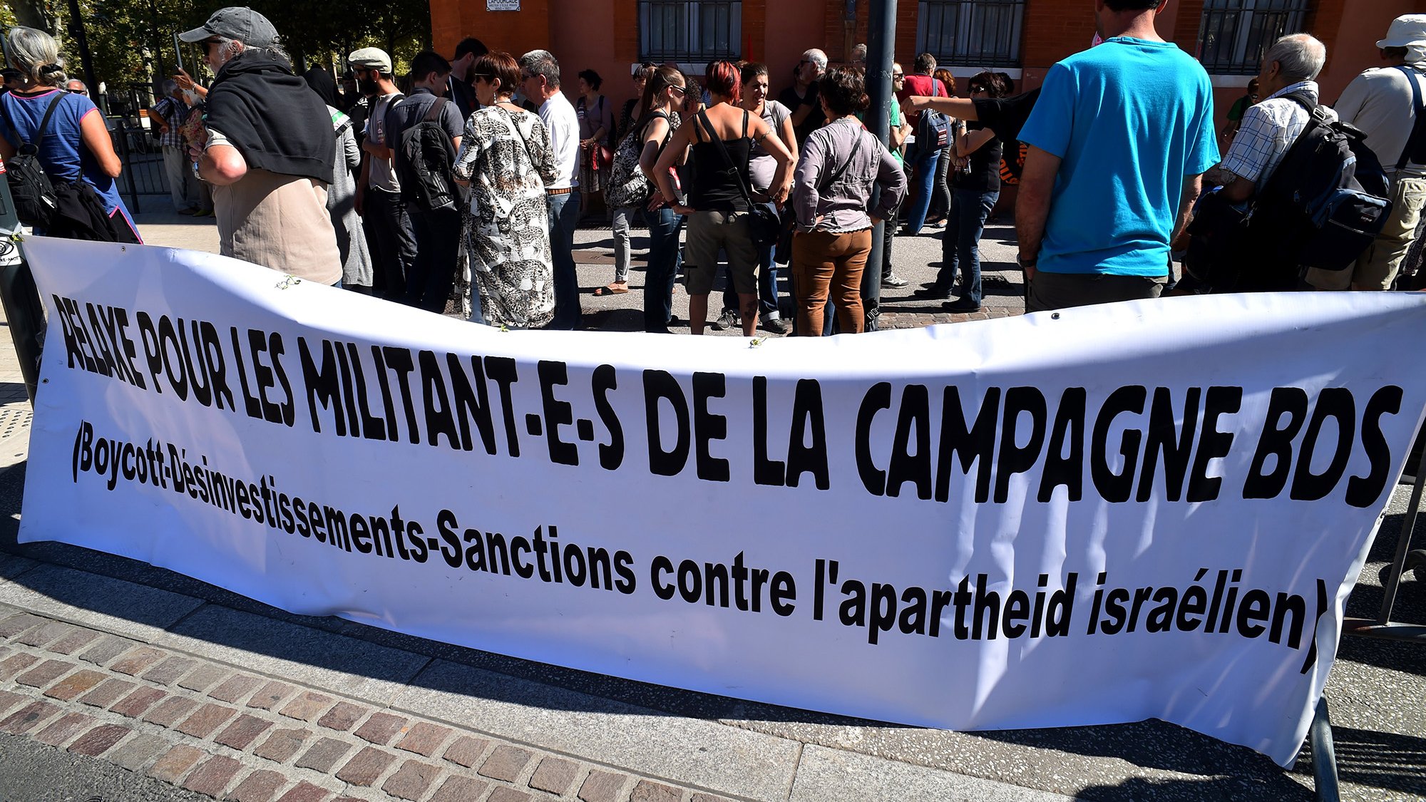 L'image montre un groupe de personnes rassemblées devant une bannière. La bannière, qui est en français, demande un soutien pour les militants de la campagne BDS, qui signifie Boycott, Désinvestissement et Sanctions contre l'apartheid israélien. Les manifestants semblent engagés et attentifs, participant à une action de protestation. Le décor urbain en arrière-plan indique que l'événement se déroule dans une ville, probablement en plein air.