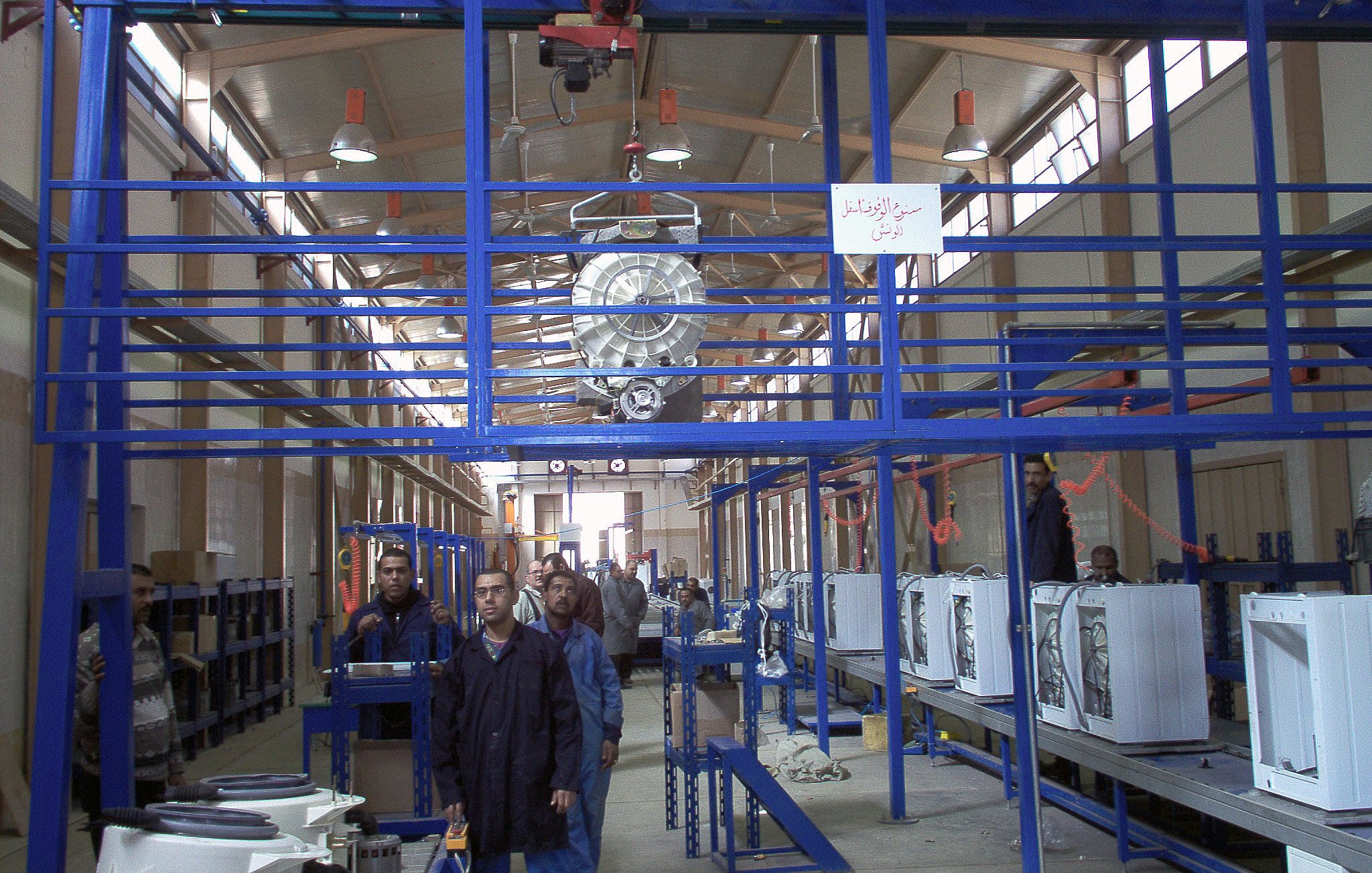 L'image montre un environnement d'atelier ou d'usine. On peut y voir des structures en métal, probablement des systèmes de rangement ou de support. Il y a des personnes travaillant dans cet espace, accompagnées d'équipements comme des appareils électroménagers, qui semblent être en phase de montage ou de préparation. L'éclairage est suffisant, et l'agencement des machines et des travailleurs suggère une ligne de production ou un processus d'assemblage. On note également des éléments rigides de la construction qui maintiennent l'espace organisé.