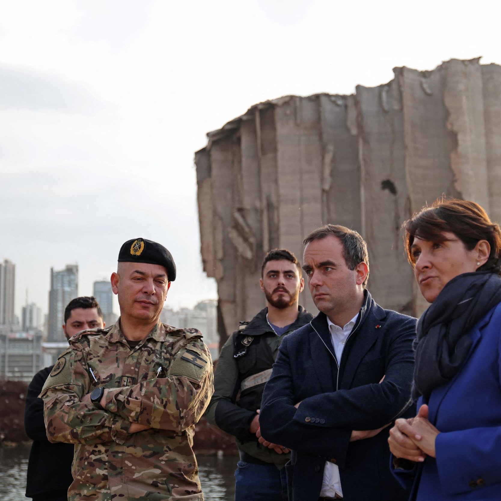 L'image montre plusieurs personnes rassemblées devant une structure endommagée, probablement un bâtiment ou un silo, avec un paysage urbain en arrière-plan. Parmi eux, on distingue un militaire en uniforme, entouré d'autres individus, dont des hommes et des femmes, portant des vêtements civils. L'ambiance semble sérieuse, comme si une discussion ou une évaluation de la situation était en cours. La lumière du jour diminue, suggérant que c'est en fin d'après-midi ou en début de soirée.
