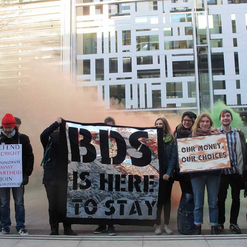 The image depicts a group of protesters standing outside a modern building, holding various signs. The signs convey messages related to support for the BDS movement, defending local democracy, and advocating for financial choices. The atmosphere seems charged, possibly with smoke or color in the background, emphasizing the protest's fervor. The group consists of diverse individuals, some wearing casual attire, and they appear to be unified in their cause.