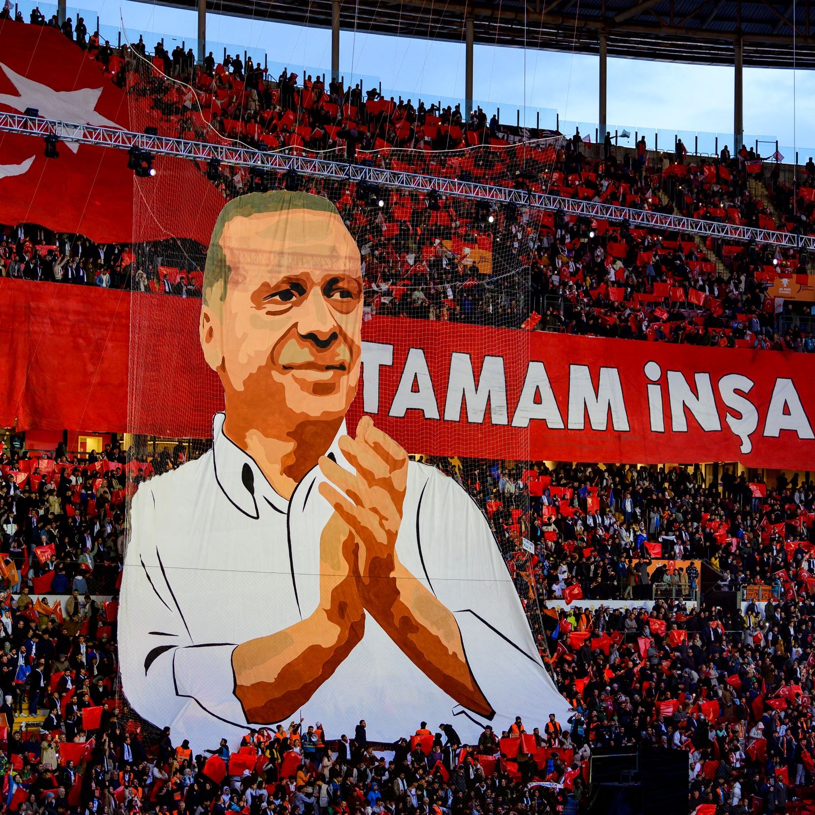 The image depicts a large crowd in a stadium, with a significant banner displayed prominently. The banner features a representation of a person, possibly a political figure, who is shown applauding. Surrounding the figure are enthusiastic supporters holding a sea of red flags or banners. The text "TAMAM İNŞALLAH" is written in bold letters, which translates to "It will be done, God willing" in English, conveying a message of hope or determination. The overall atmosphere appears vibrant and charged with energy, indicative of a political rally or celebration.