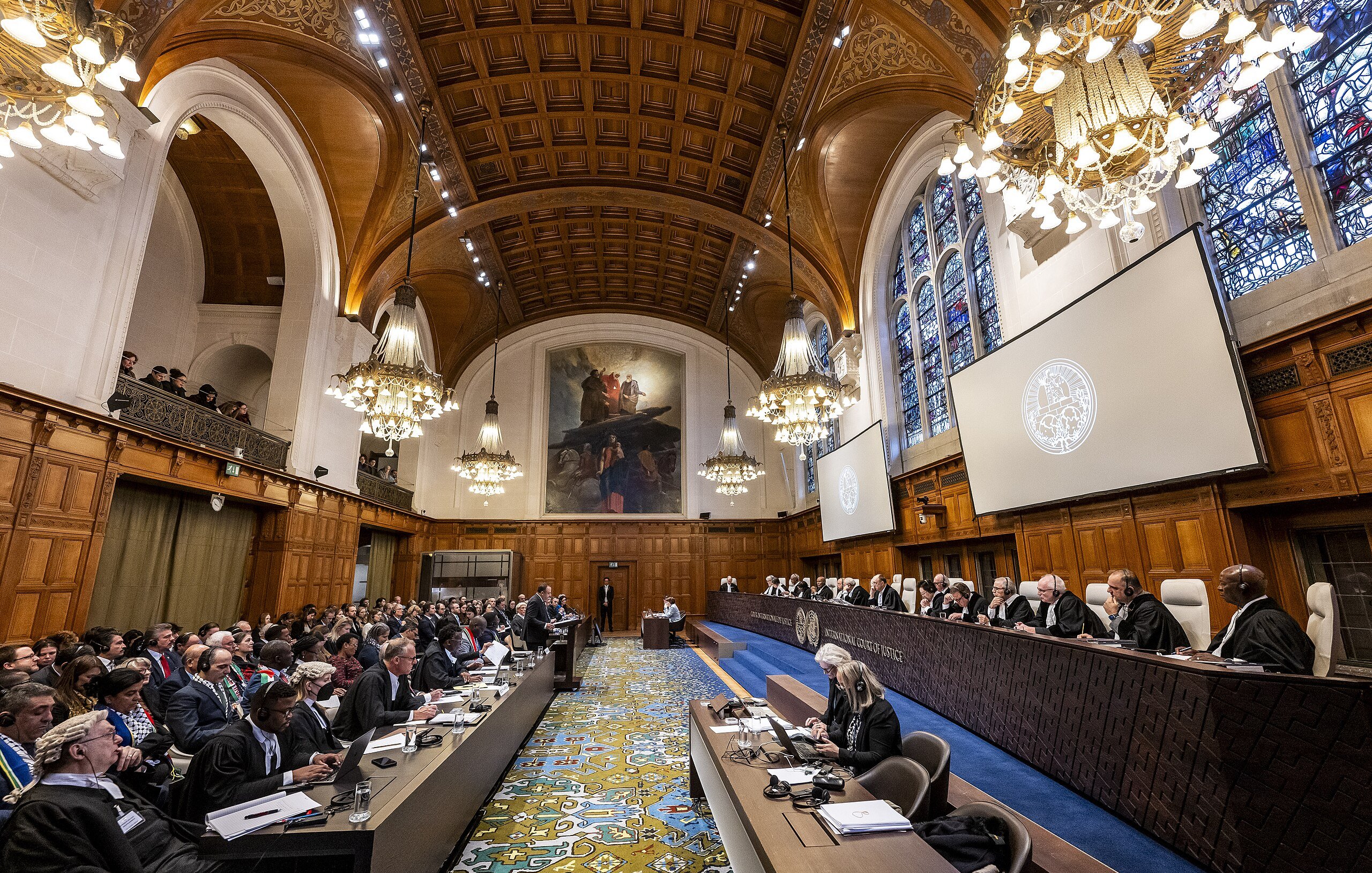 L'immagine mostra un'aula di giustizia, probabilmente una corte internazionale. La sala è caratterizzata da un'architettura imponente con soffitti alti, lampadari eleganti e finestre ad arco che lasciano filtrare la luce. In primo piano, c'è un lungo tavolo con diversi avvocati e rappresentanti, mentre sul lato opposto, una serie di giudici sono seduti su una piattaforma rialzata. Il pubblico è composto da molte persone, alcune delle quali prendono appunti. L'atmosfera sembra formale e rispettosa, tipica di un'importante udienza legale.