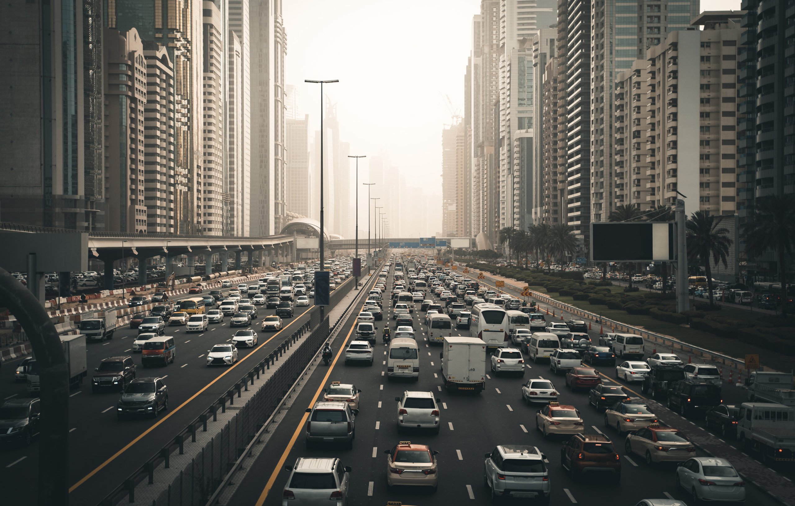 L'image montre une vue d'une route très fréquentée dans une grande ville moderne, entourée de gratte-ciel. On peut voir de nombreux véhicules qui circulent dans les deux sens, créant un embouteillage. La lumière du soleil est tamisée, donnant une atmosphère légèrement brumeuse, peut-être due à la pollution ou à la météo. Les bâtiments en arrière-plan sont imposants et reflètent l'architecture contemporaine, avec des façades en verre et en béton. Des palmiers sont visibles sur le côté de la route, ajoutant une touche de verdure à ce paysage urbain.