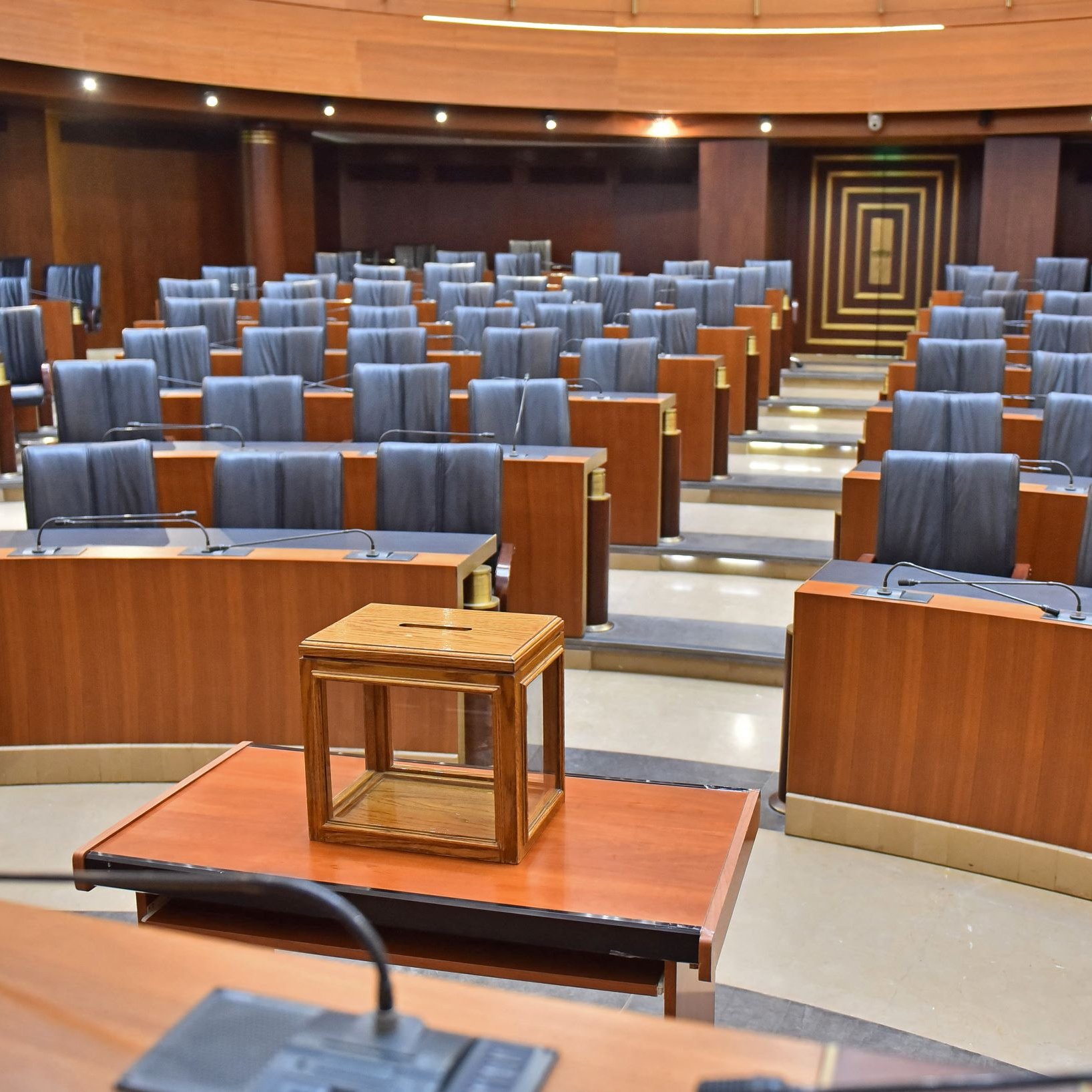 L'immagine mostra un'aula legislativa vuota, caratterizzata da una disposizione ordinata di banchi in legno con sedute nere. Al centro della scena si trova una piccola struttura in legno, probabilmente un tavolo o una cassa. Le pareti dell'aula sono di un colore scuro, mentre il pavimento è di tonalità più chiara, creando un contrasto. L'ambiente sembra formale e istituzionale, suggerendo un luogo di discussione e decisione politica.