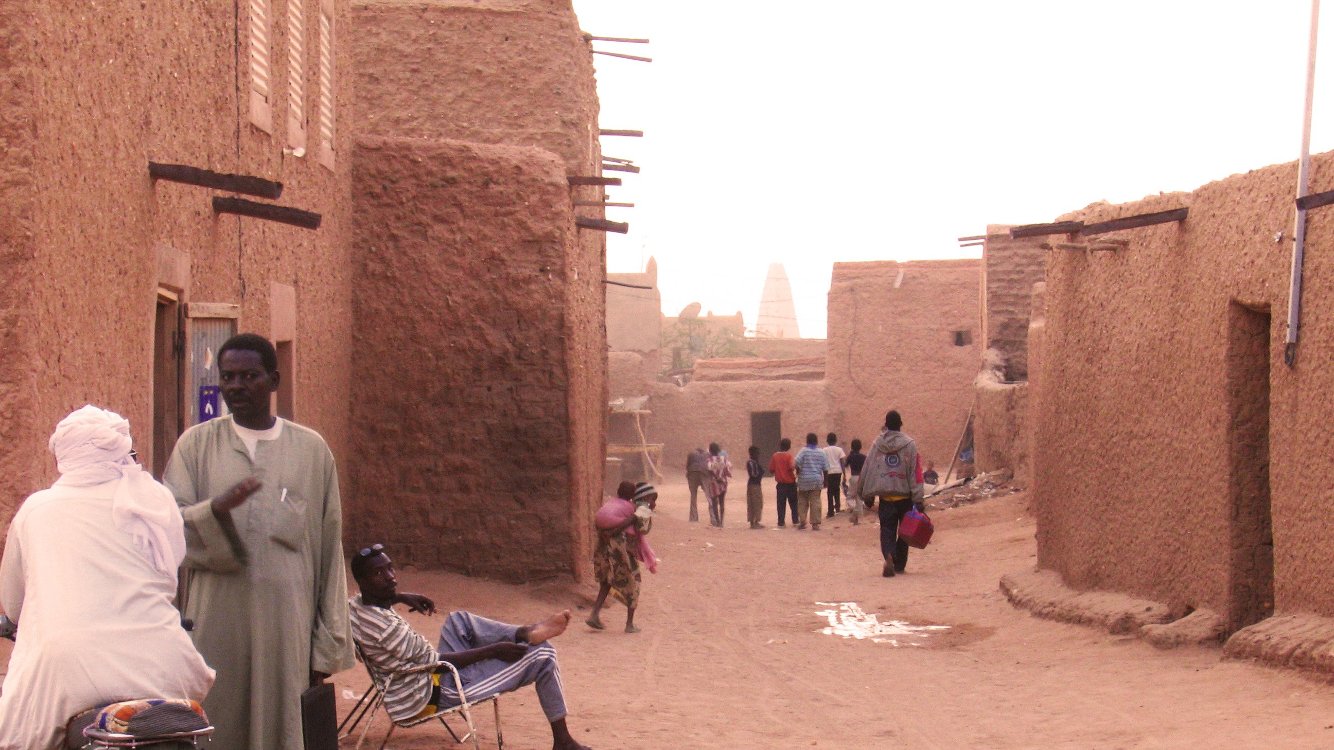 L'image montre une rue dans un village traditionnel, probablement situé dans une région désertique. On y voit des maisons en terre, avec des murs de couleur ocre. Plusieurs personnes déambulent tranquillement dans la rue, certaines s'arrêtent pour discuter. À gauche, un homme est assis sur une moto, tandis qu'un autre se repose sur une chaise. L'atmosphère est calme et chaleureuse, avec un éclairage doux peut-être dû au coucher du soleil. Les ombres et la poussière soulignent le caractère rustique du lieu.