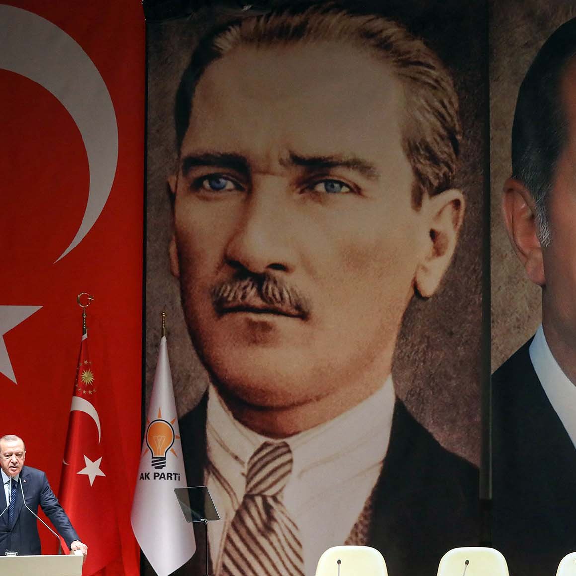 L'immagine ritrae un evento politico in Turchia. Sullo sfondo, sono presenti due grandi ritratti: uno di Mustafa Kemal Atatürk, fondatore della Repubblica turca, e l'altro di Recep Tayyip Erdoğan, attuale presidente. Al centro, un oratore sembra parlare al pubblico, con bandiere turche e simboli del partito sul palco. Lo sfondo con i due leader storici suggerisce un legame tra il passato e il presente politico del paese.