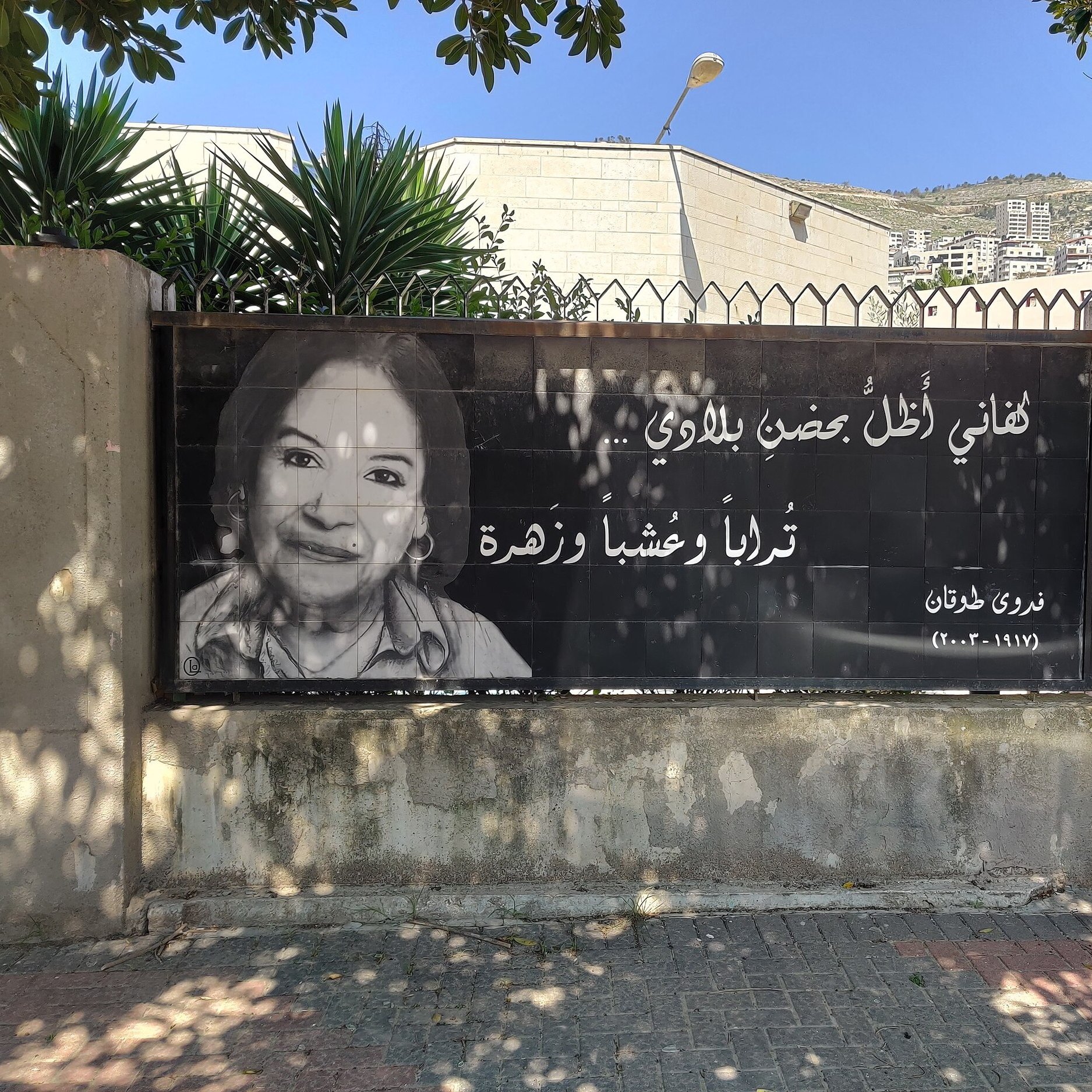 L'immagine mostra un grande striscione che riporta una fotografia di una donna sorridente, accompagnata da un testo in arabo. Il contesto sembra essere un'area urbana, con edifici sullo sfondo e piante verdi nelle vicinanze. L'atmosfera è accogliente e il messaggio sullo striscione appare positivo e invitante.