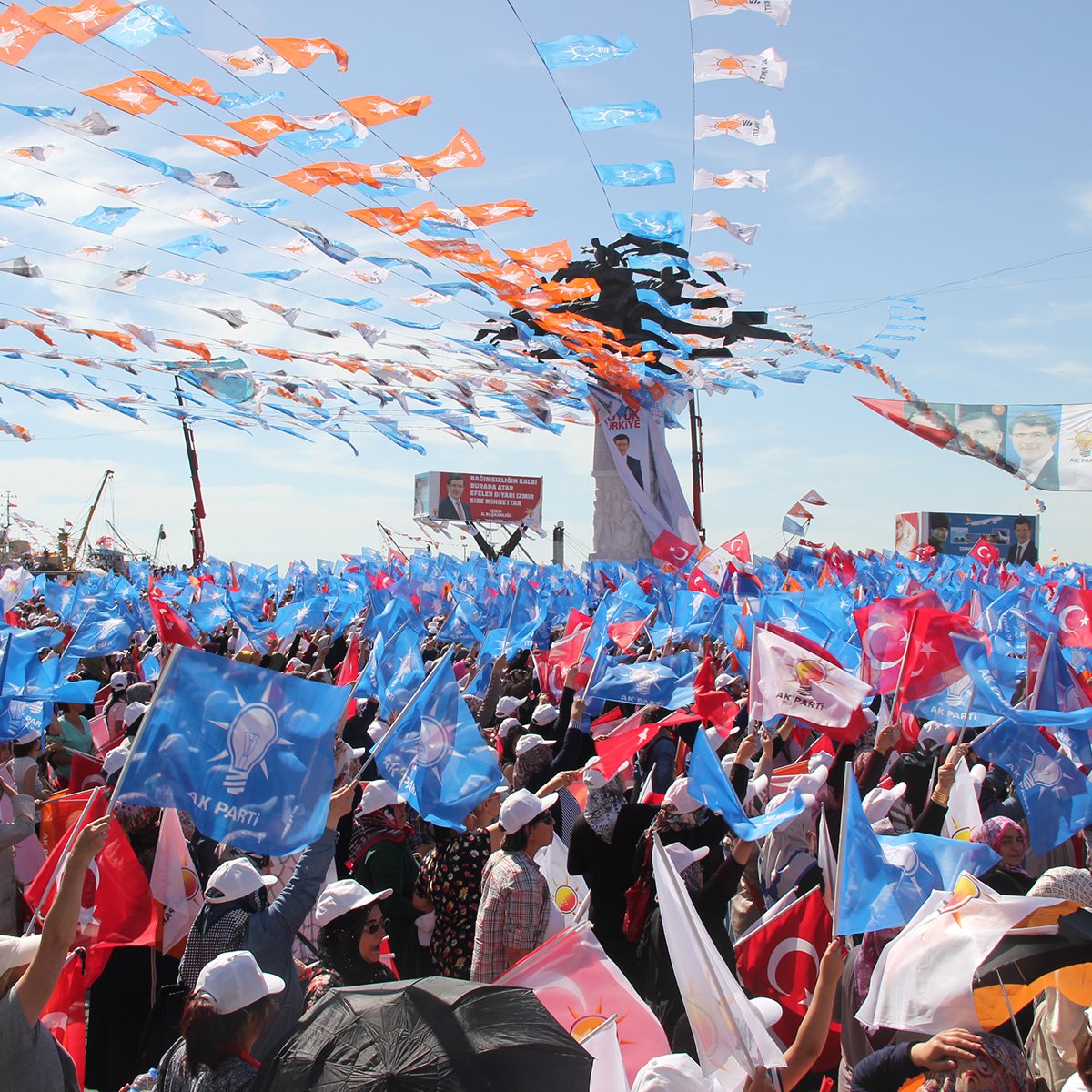 The image depicts a large gathering or rally, filled with a sea of people holding colorful flags. The flags appear to be in various shades of blue, orange, and other colors, creating a vibrant atmosphere. Many attendees are wearing white hats and there are banners or signs visible in the background. The sky is bright, suggesting a sunny day, and the scene conveys a sense of energy and excitement among the crowd.