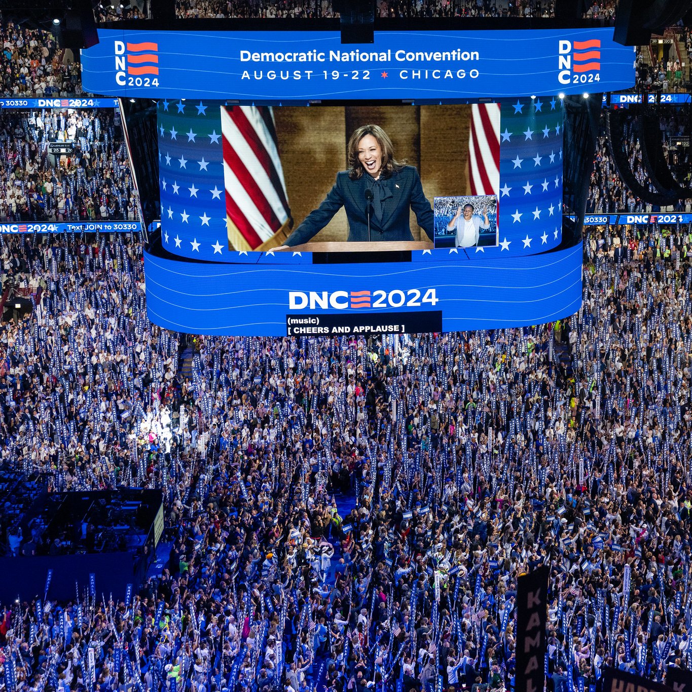 این تصویر نمایی از کنوانسیون ملی دموکرات‌ها در سال 2024 را نشان می‌دهد. در مرکز تصویر، یک نفر روی سن ایستاده و به جمعیت حاضر در سالن خطاب می‌کند. پس‌زمینه شامل پرچم‌های دموکراتیک و تبلیغات مربوط به کنوانسیون است. جمعیت زیادی در سالن حضور دارند و برخی از آن‌ها تابلوهایی در دست دارند. نورپردازی و طراحی صحنه به وضوح توجهات را جلب کرده و جو پرشوری را به تصویر می‌کشد. تاریخ و محل برگزاری کنوانسیون نیز در بالای تصویر نوشته شده است.