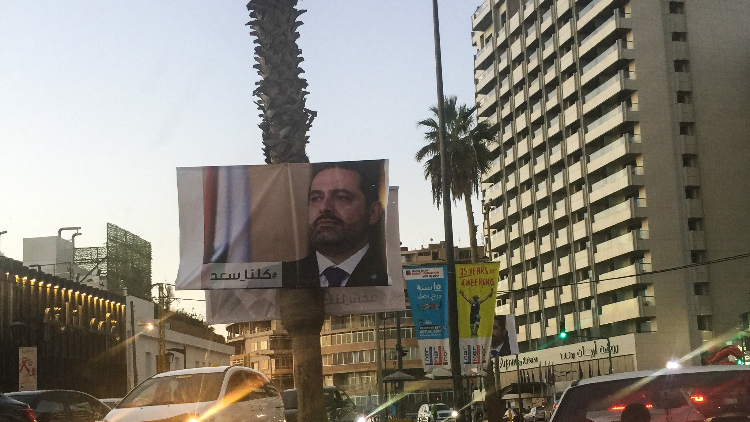 در این تصویر یک بیلبورد بزرگ در کنار خیابان دیده می‌شود که چهره یک مرد را نمایش می‌دهد. او نسبت به دوربین نگاهی جدی دارد و به نظر می‌رسد که یک شخصیت سیاسی باشد. در زمینه، ساختمان‌های بلند و درختان نخل دیده می‌شوند که فضای شهری را تشکیل می‌دهند. نور غروب خورشید بر روی صحنه افکنده شده که به آن حال و هوای خاصی می‌بخشد. چندین خودرو نیز در خط خیابان در حال حرکت هستند.