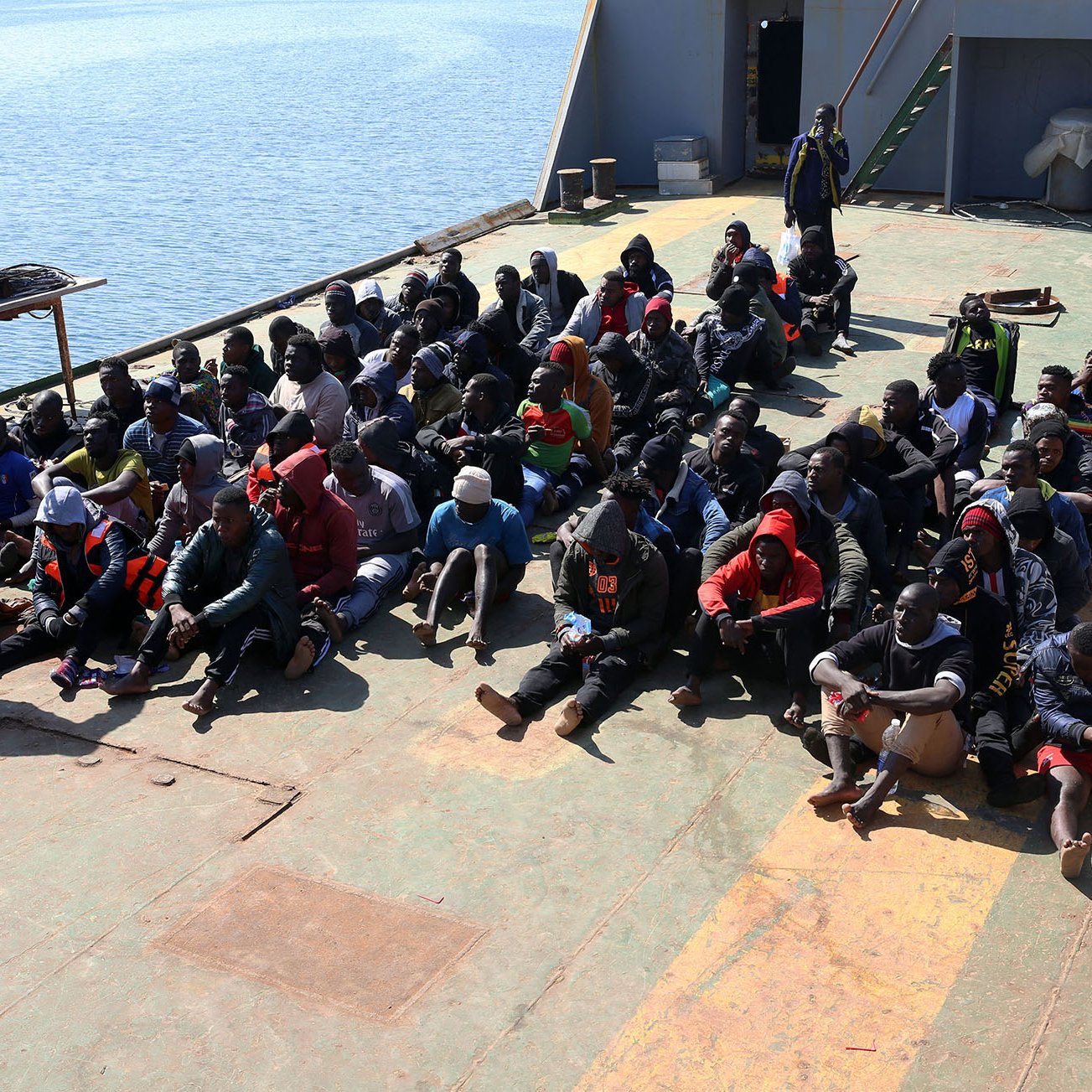 در این تصویر، گروهی از افراد در یک کشتی نشسته‌اند. آنها در حالتی متراکم و منظم روی عرشه نشسته‌اند و بیشتر آنها لباس‌های گرم به تن دارند. برخی از آنها هم کلاه و ماسک به صورت دارند. در سمت چپ تصویر، فردی با یونیفورم نظامی و شخصی دیگر با جلیقه آبی در حال نظارت بر این گروه هستند. در پس‌زمینه، آب دریا و بخشی از ساحل قابل مشاهده است. جو کلی تصویر به نظر می‌رسد که مربوط به یک عملیات نجات یا مهاجرت باشد.
