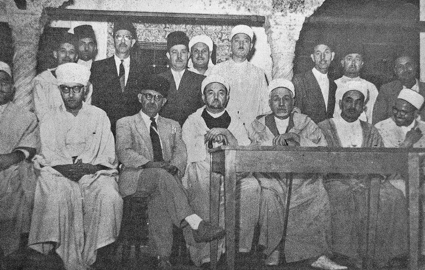 L'image montre un groupe de personnes rassemblées dans une pièce. Elles portent des vêtements traditionnels, notamment des turbans et des robes longues. Au centre, quelques individus sont assis à une table, tandis que d'autres se tiennent debout derrière eux. L'atmosphère semble formelle et solennelle, suggérant une réunion ou une cérémonie. Les murs sont en pierre, ce qui donne un aspect historique à l'ensemble.