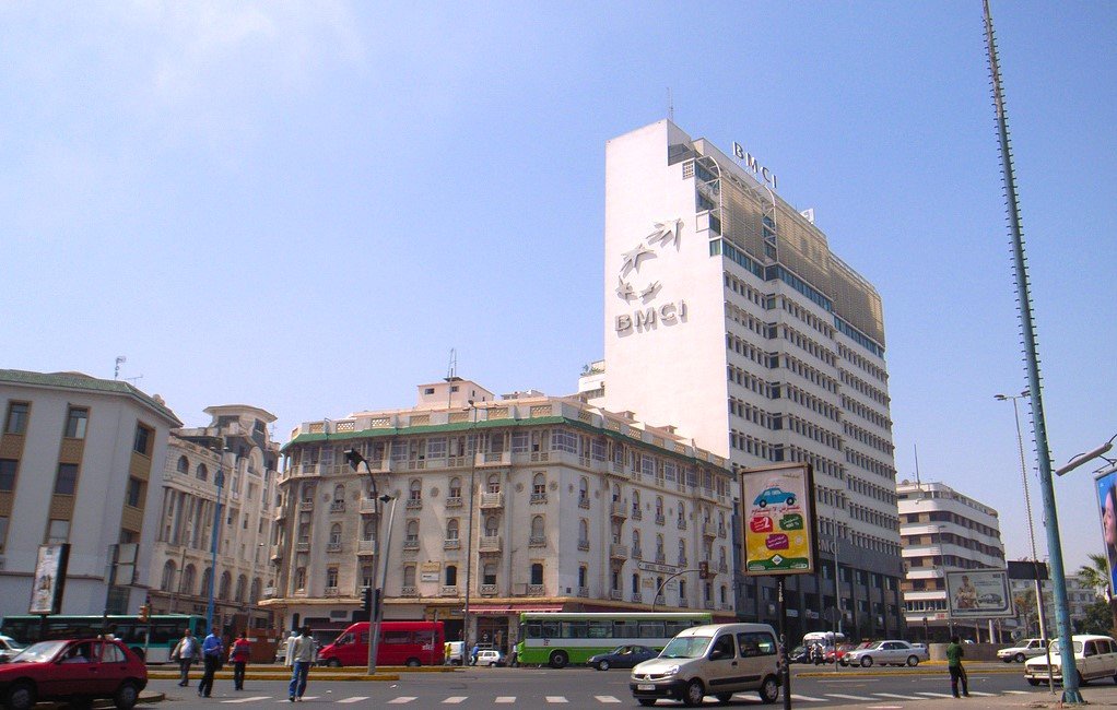 The image shows a cityscape featuring a tall building with the letters "BMCI" prominently displayed on it. The architecture of the surrounding buildings is a mix of modern and traditional styles. The sky is clear and blue, indicating a sunny day. In the foreground, there are vehicles on the road, including a red car and a few buses, as well as a billboard advertising various products. The scene conveys a bustling urban environment.