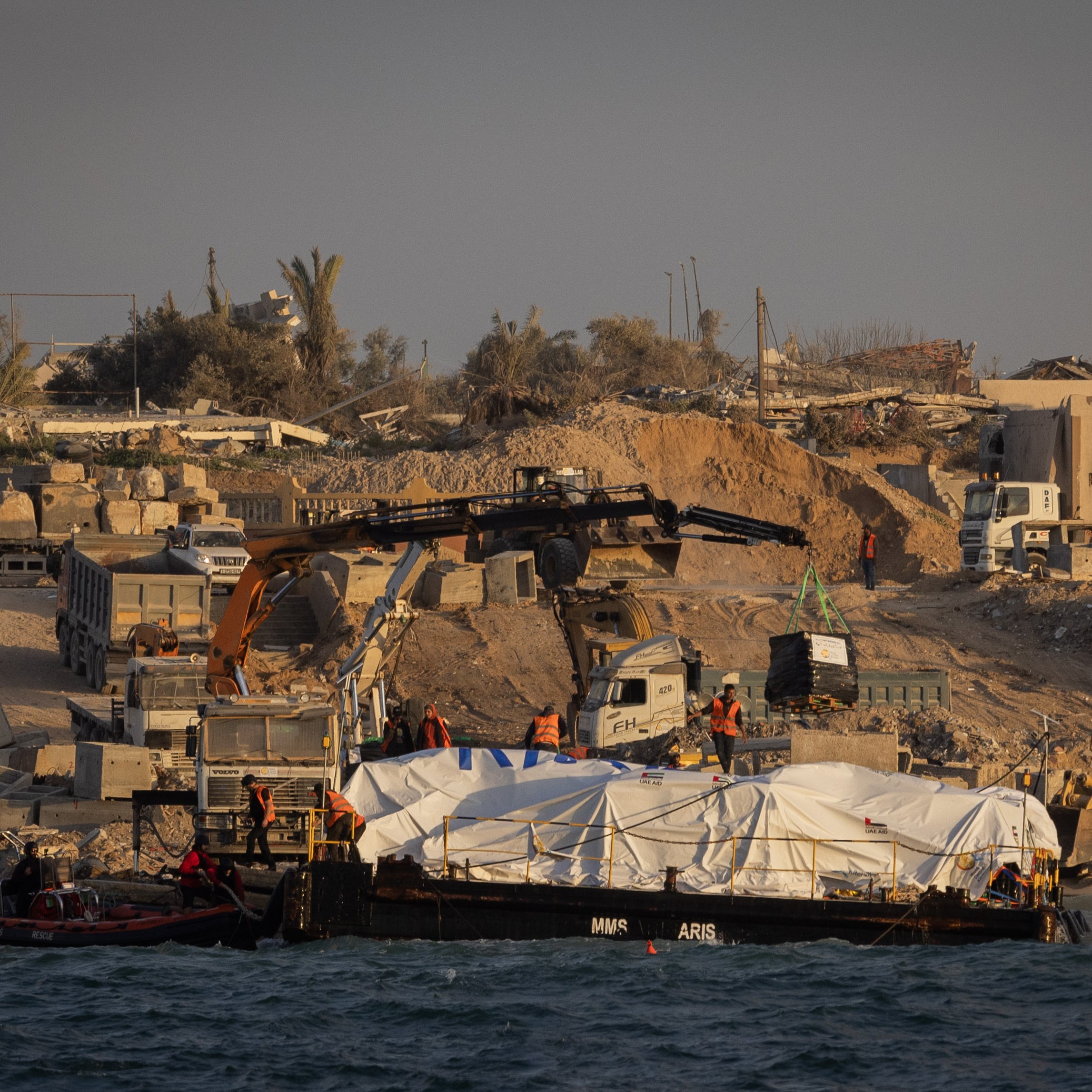 L'immagine mostra una scena costiera con un'imbarcazione che trasporta un carico coperto da un telo bianco. Sulla riva, si possono vedere mezzi di costruzione e operai al lavoro, circondati da detriti e strutture in fase di demolizione o costruzione. Sullo sfondo, ci sono alberi e edifici, con l'atmosfera tipica di un'area in fase di riqualificazione o ricostruzione. La luce del sole sembra stia calando, creando un'atmosfera suggestiva.