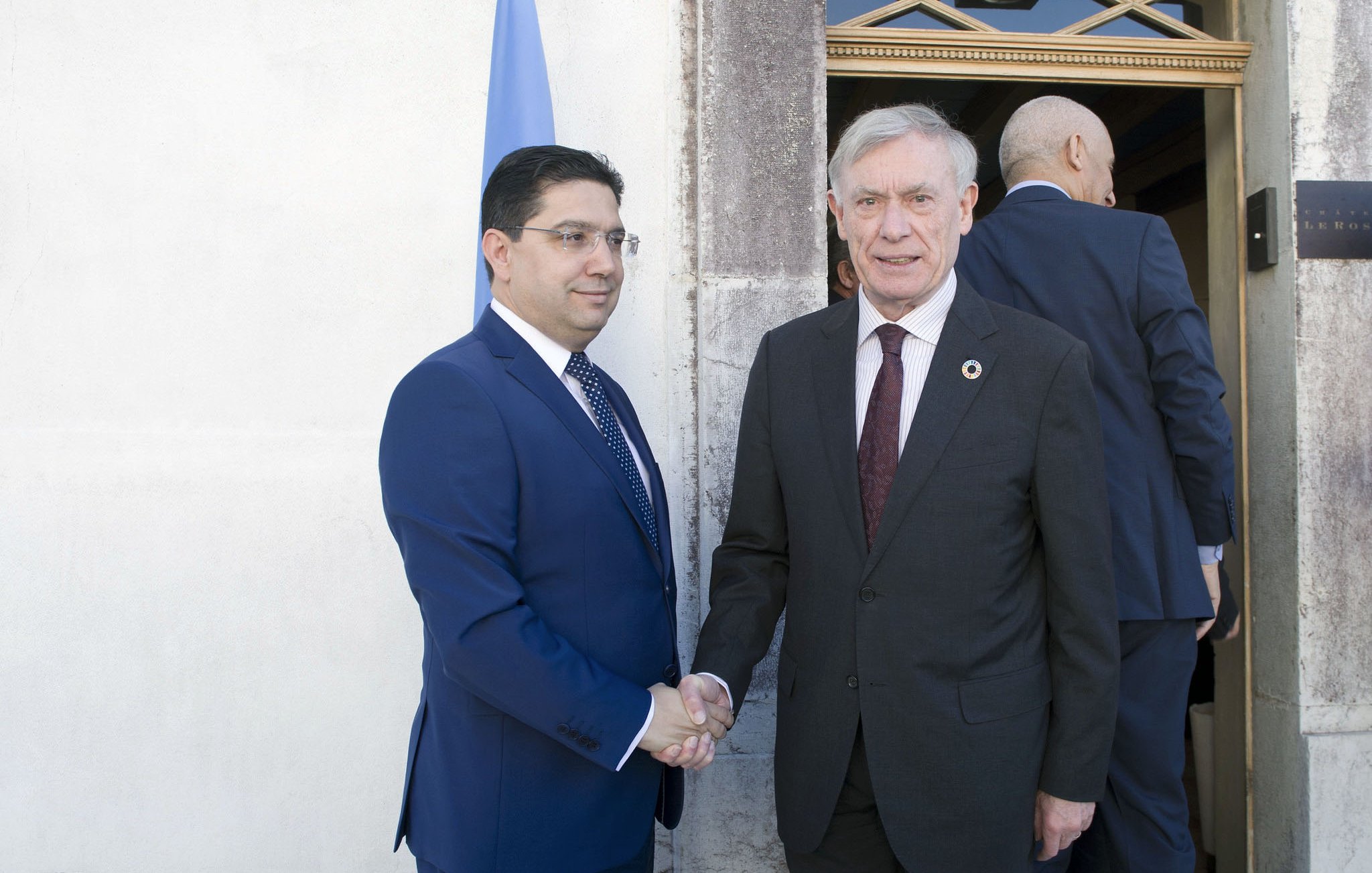 En la imagen se puede ver a dos hombres en un ambiente formal. Uno de ellos está vestido con un traje oscuro y una corbata, mientras que el otro lleva un traje azul. Ambos están dándose la mano, lo que sugiere un saludo o un acuerdo. En el fondo hay una puerta y una bandera, lo que indica que están en un entorno institucional o diplomático. La escena transmite un sentido de cordialidad y oficialidad.