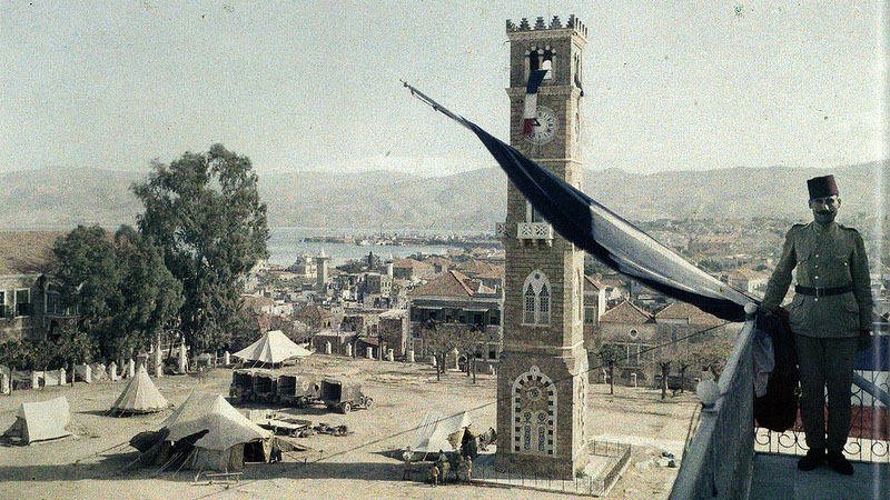 L'image montre une vue panoramique d'une place animée avec un grand bâtiment en pierre qui pourrait être une horloge ou une tour emblématique. À proximité, un soldat en uniforme se tient près de la balustrade, observant la scène. On peut voir des tentes disposées sur la place, peut-être pour un camp militaire ou une exposition. En arrière-plan, on distingue des collines et un plan d'eau, ajoutant à la profondeur du paysage. Le ciel semble dégagé, créant une atmosphère paisible.