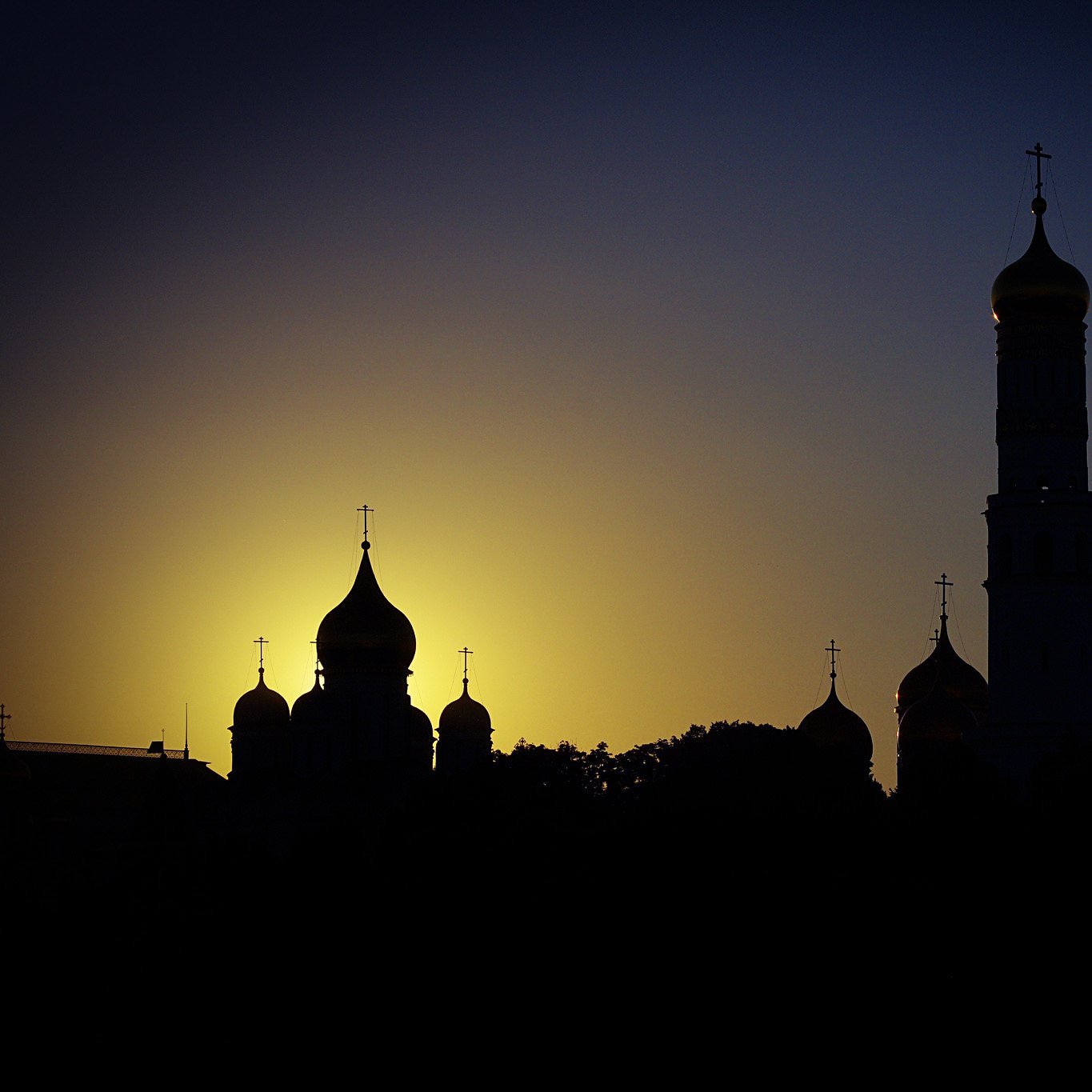 The image features a silhouette of a skyline, likely of a historic city or religious site, during sunset. In the foreground, various domes and towers with crosses are visible, indicating the presence of churches or cathedrals. The background is illuminated by the warm glow of the setting sun, creating a gradient from dark blue to golden yellow. The overall atmosphere suggests tranquility and reverence, highlighting the architectural beauty against the fading light.