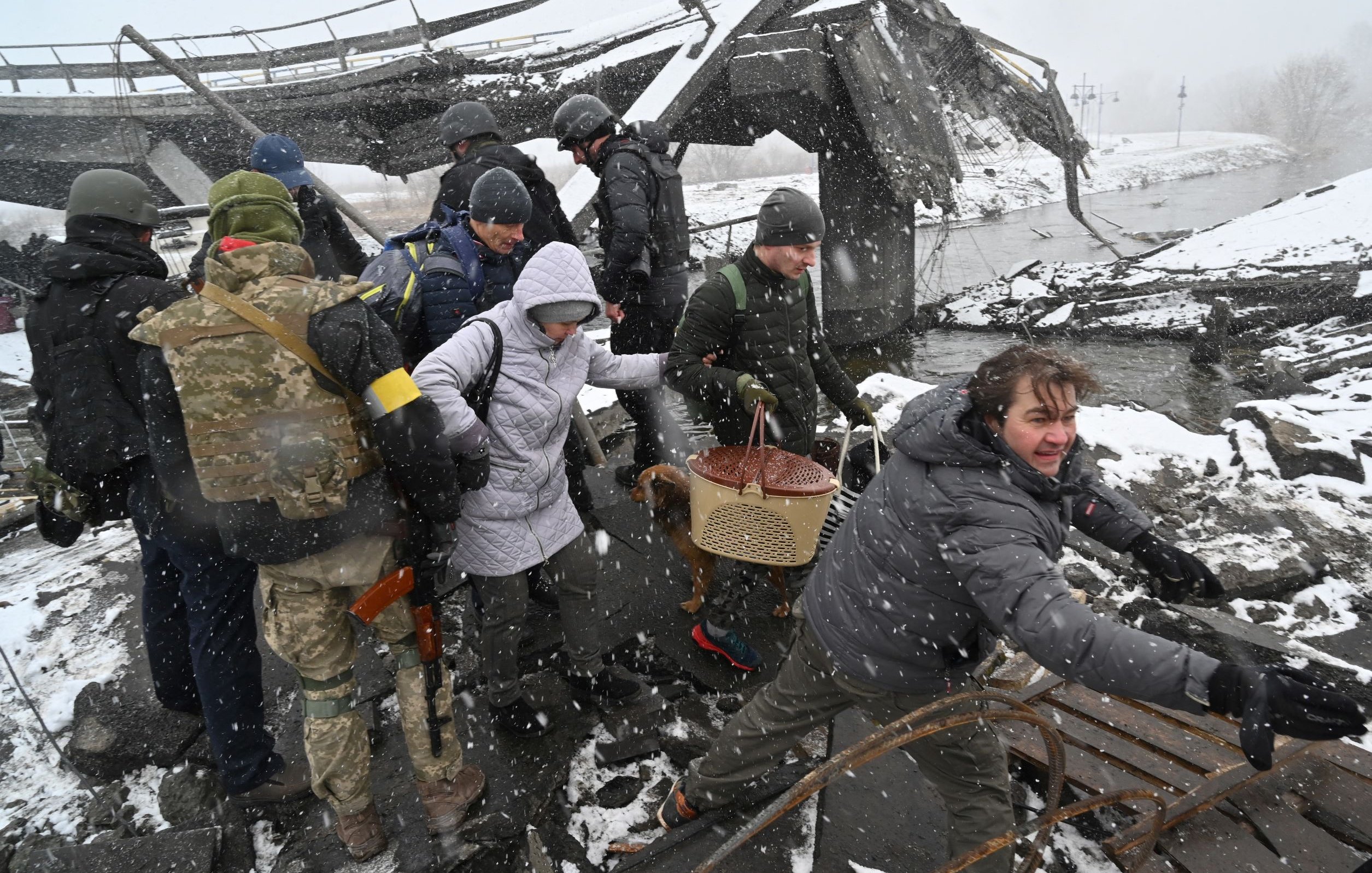 The image depicts a group of people navigating through a damaged area, likely following a disaster or conflict. They are surrounded by debris and snow, suggesting challenging weather conditions. Some individuals appear to be assisting others, while one person is pulling or reaching for something. The scene conveys a sense of urgency and struggle in a harsh environment, highlighting the effects of destruction and the resilience of those affected.