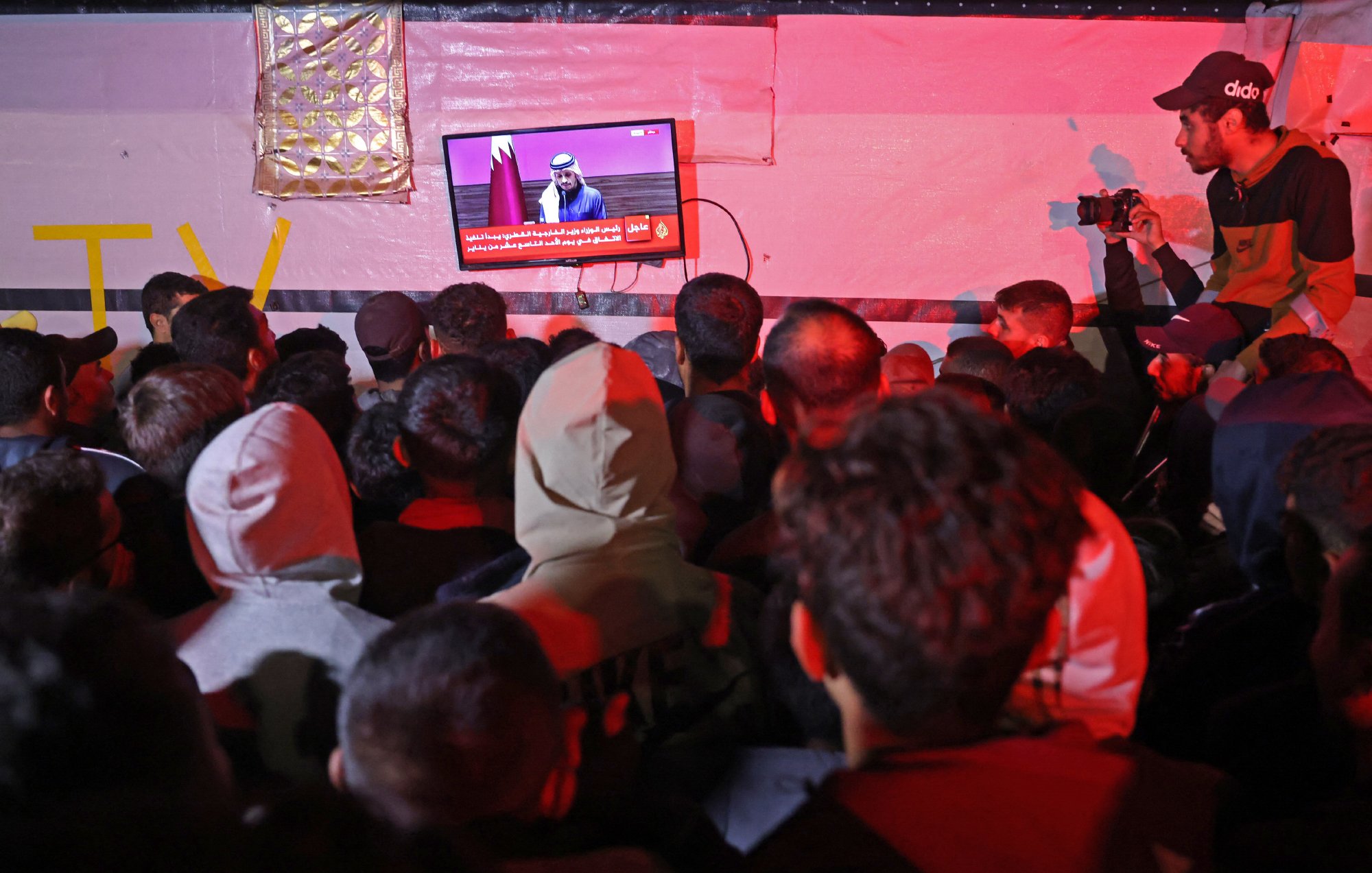 L'image montre une foule de personnes rassemblées, observant attentivement un écran au-dessus d'eux. L'écran affiche un discours ou une annonce, probablement en rapport avec des événements d'actualité. Certaines personnes dans la foule portent des capuches et des vêtements chauds, ce qui suggère qu'il fait frais. Un individu à droite prend des photos, ajoutant une dimension journalistique à la scène. L'éclairage est tamisé, créant une ambiance particulière.