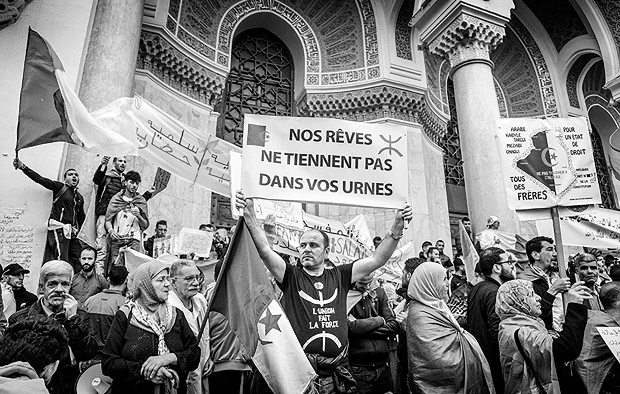 L'immagine ritrae una manifestazione di protesta. Al centro, un uomo tiene in alto un cartello con scritto "NOS RÊVES NE TIENNENT PAS DANS VOS URNES", che significa "I nostri sogni non stanno nelle vostre urne". Sullo sfondo, ci sono altri manifestanti e striscioni recanti vari messaggi. L'atmosfera sembra intensa, con persone che esprimono il loro dissenso e la loro determinazione. L'immagine è in bianco e nero, aggiungendo un senso di drammaticità e urgenza alla scena.