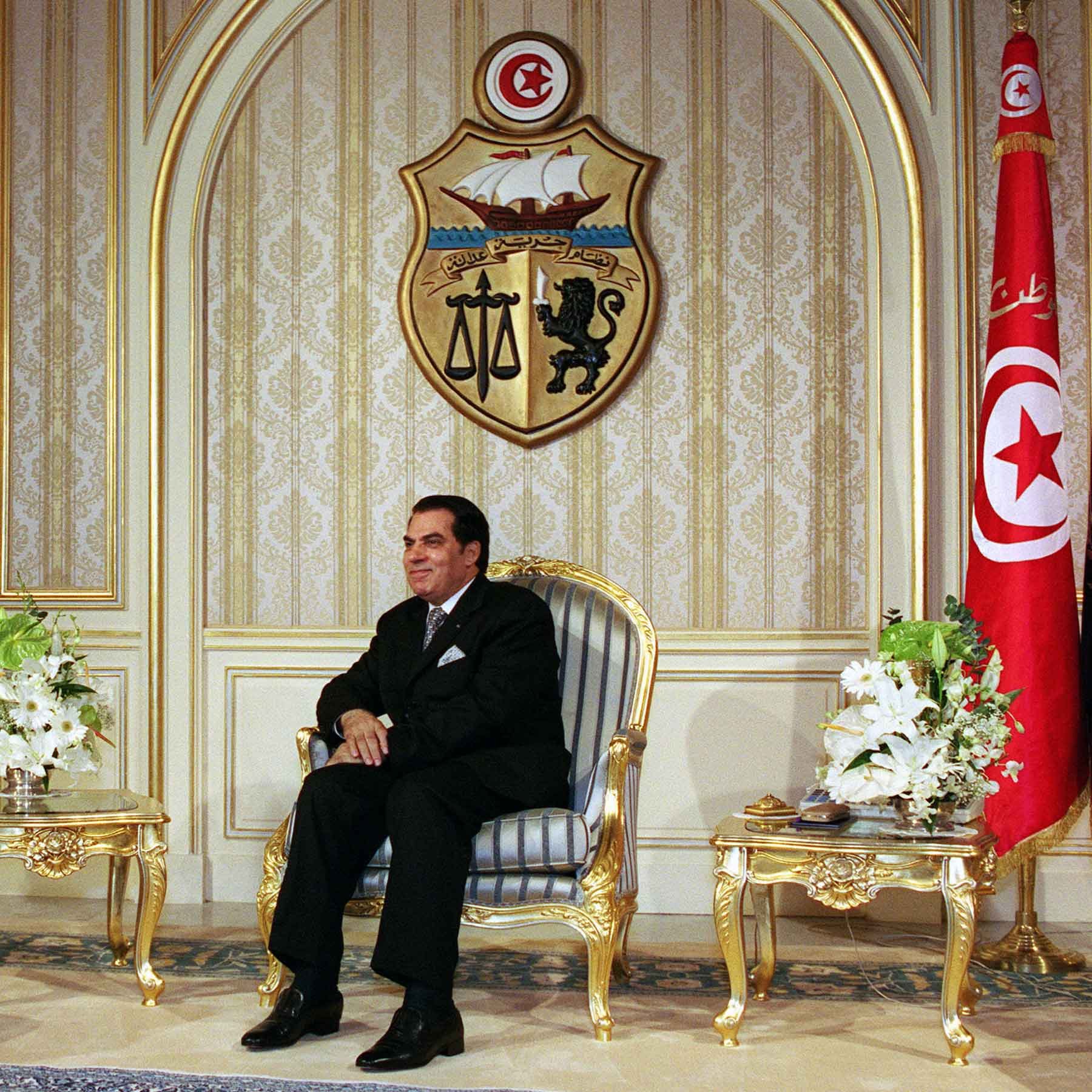 L'image montre un homme assis sur une chaise élégante dans un environnement formel. Il porte un costume sombre et semble très confortable. À l'arrière-plan, on peut voir un grand blason tunisien ainsi qu'un drapeau de la Tunisie, ce qui indique qu'il s'agit d'un cadre officiel. Des fleurs blanches sont placées sur des tables basses de chaque côté de lui, ajoutant une touche de sophistication à la scène. Les murs sont décorés de manière riche, ce qui renforce le caractère solennel de l'environnement.