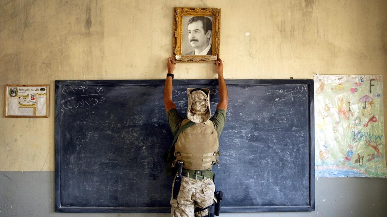 L'image montre un soldat en uniforme, portant un gilet de protection, en train de fixer un portrait dans un cadre doré sur un mur. Le portrait représente un homme aux traits marqués, probablement un dirigeant. En arrière-plan, on peut voir un tableau noir avec des inscriptions en arabe, ainsi qu'une feuille de papier accrochée au mur, qui semble être un dessin. L'atmosphère de la scène semble refléter un environnement qui a été, ou est, lié à des événements militaires ou politiques.