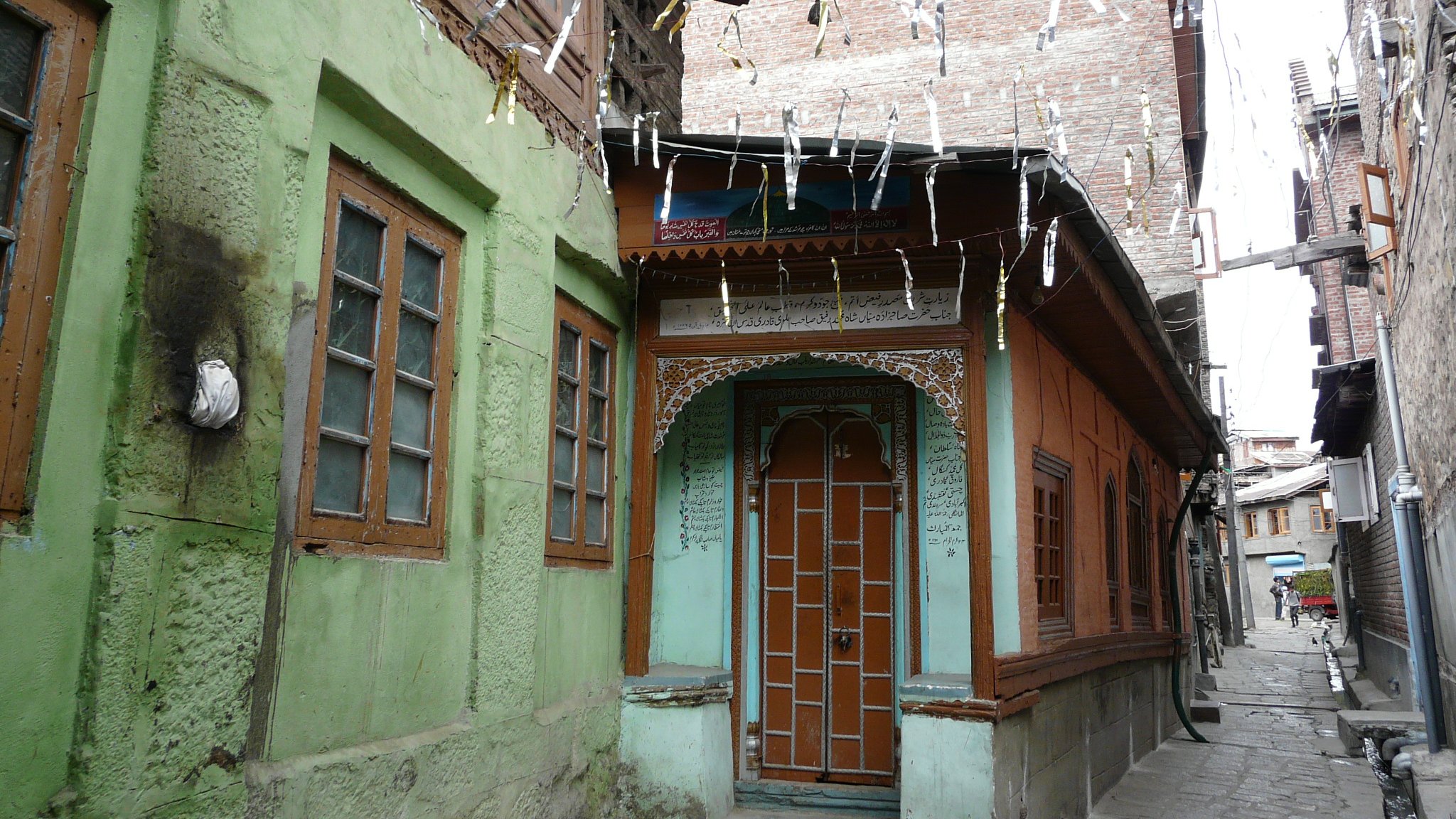 L'image montre une rue étroite et pittoresque dans une vieille ville, avec des maisons en bois et en pierre. On peut voir des façades colorées, notamment des murs verts et oranges, ainsi que des fenêtres à volets en bois. Les bâtiments ont un style architectural traditionnel, avec des détails ornementaux visibles. Des fils sont tendus entre les maisons, probablement pour des décorations ou des lumières. L'atmosphère évoque un sentiment de nostalgie et de charme ancien.