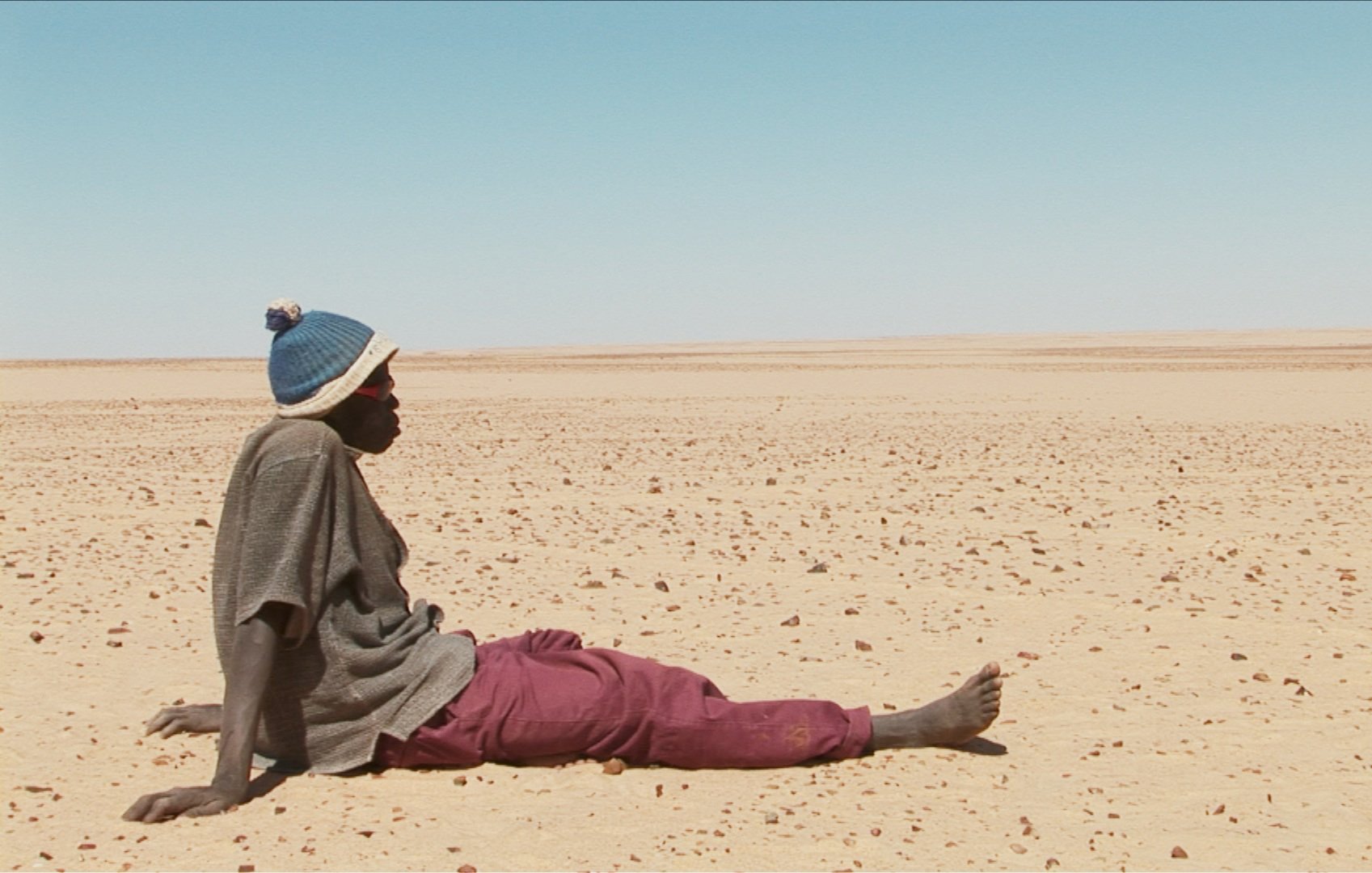 L'image montre un homme assis sur le sol désertique. Il porte un pull avec un bonnet et un pantalon de couleur prune. Le paysage autour de lui est vaste et aride, constitué principalement de sable et de petites pierres, s'étendant à perte de vue sous un ciel clair. L'atmosphère semble à la fois isolée et silencieuse, évoquant une sensation de solitude dans cet environnement désertique.