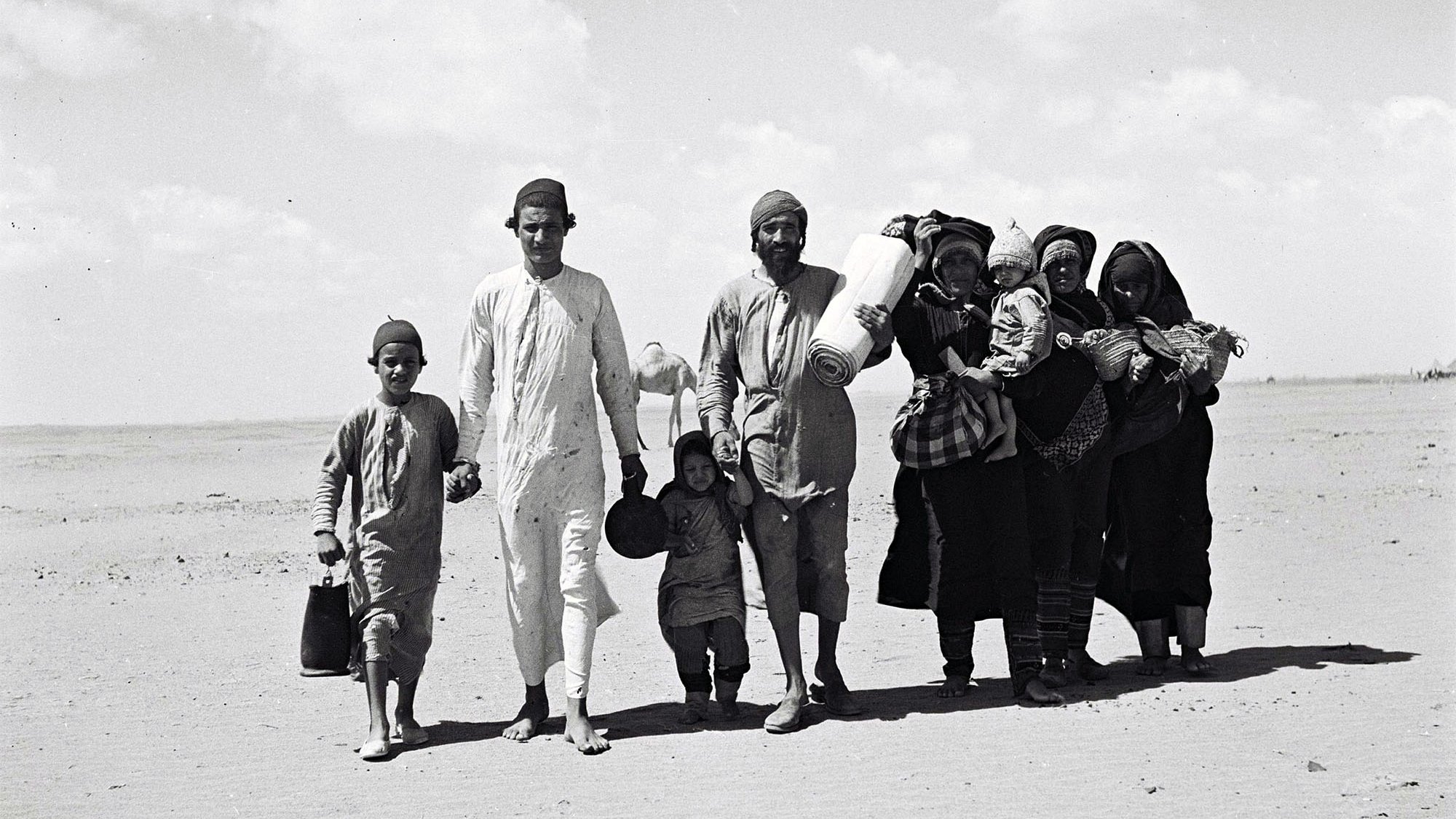 The image depicts a group of people walking across a sandy landscape. There are five adults and three children, with some carrying bundles and containers. The adults wear traditional clothing, and the children are dressed in simpler attire. The setting appears to be arid, with a clear sky and sparse vegetation in the background. The group's expressions seem serious as they move forward, suggesting a journey or migration.