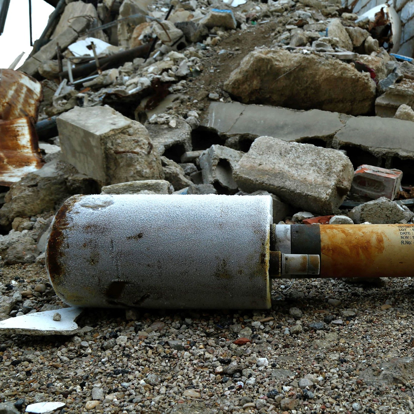 The image shows a missile or bomb lying on the ground among rubble and debris. The surrounding area is chaotic, with broken concrete and scattered materials suggesting destruction, possibly from an explosion or military action. The missile itself appears weathered and is partially covered in grime, indicating it has been in this location for some time.