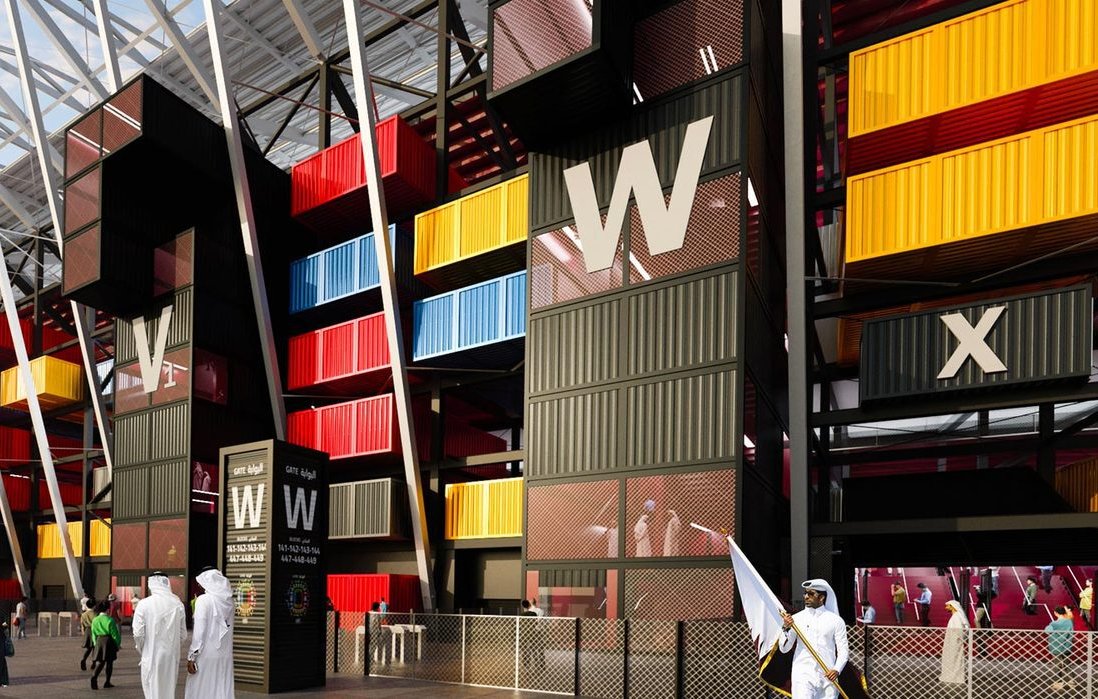 The image depicts a modern architectural structure that resembles a stadium or a large public venue. The building features a striking façade made up of brightly colored shipping containers in red, yellow, and blue, arranged in a visually dynamic way. There are tall, angular elements with a white roof framework above. In the foreground, a group of people dressed in traditional attire can be seen, indicating a cultural context. Overall, the design conveys a sense of contemporary style and innovative use of space.