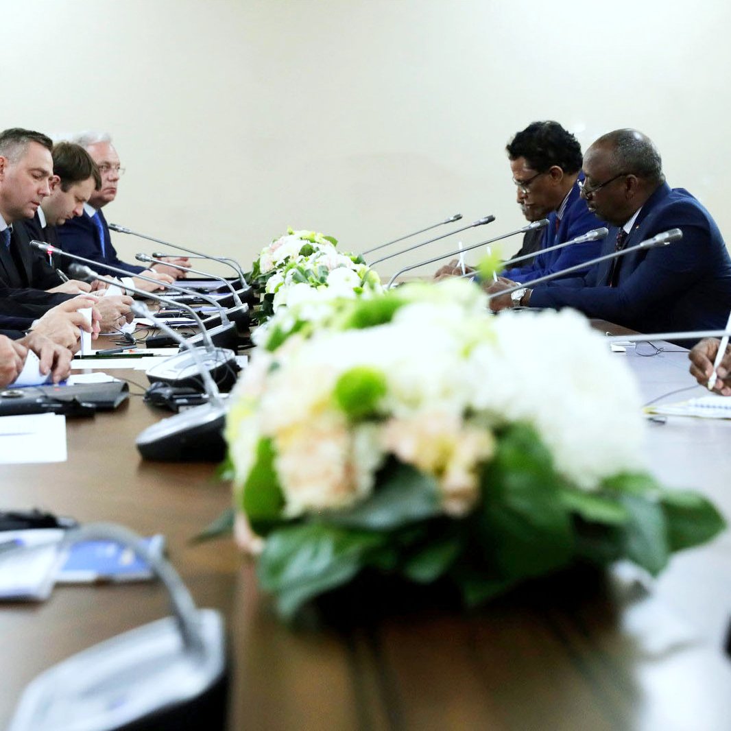 The image depicts a formal meeting or conference, with a long table where several individuals are seated across from each other. There appears to be a floral centerpiece on the table. The participants seem to be engaged in discussion or note-taking, with some looking attentively at documents or each other. The setting appears to be a neutral, professional environment, likely a government or diplomatic meeting.