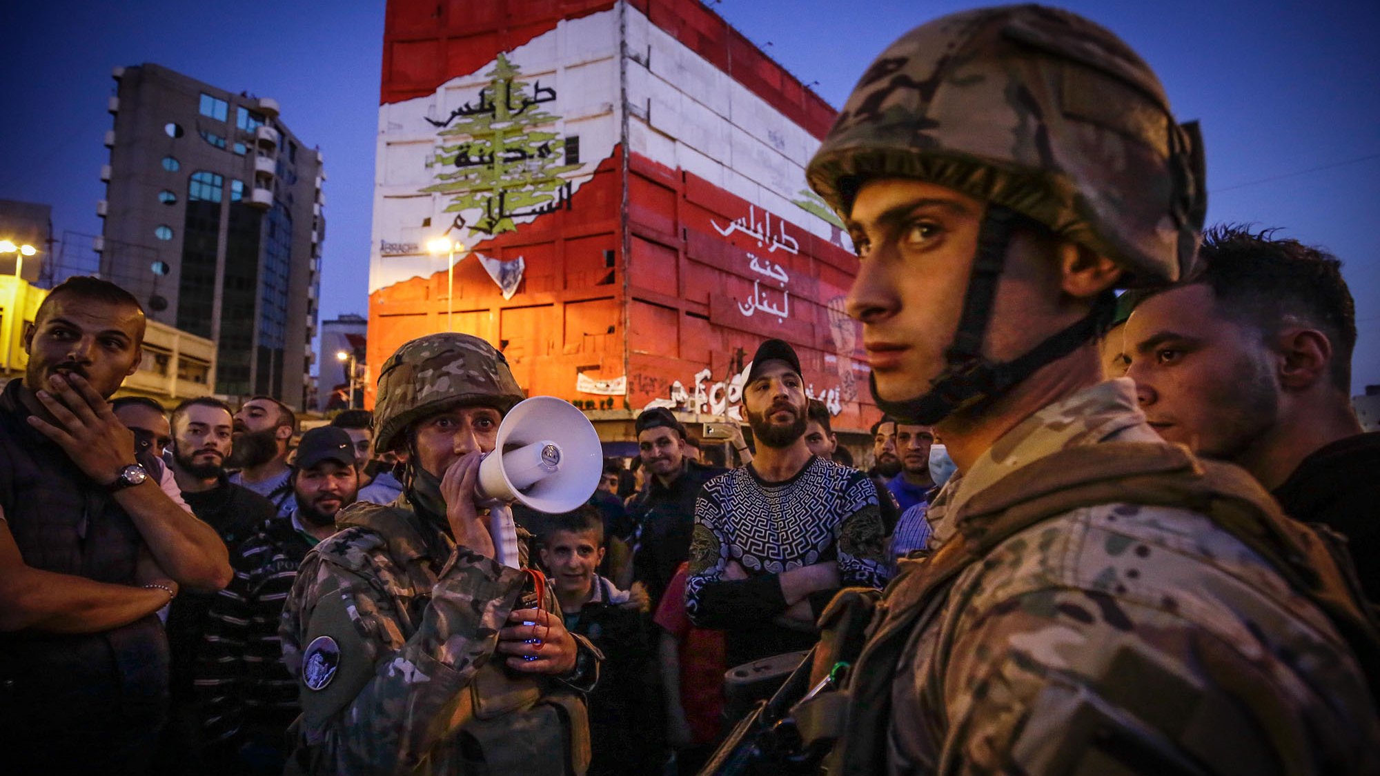 The image depicts a scene of a gathering in an urban environment during the evening. In the foreground, soldiers in military gear, including helmets, are present, with one holding a megaphone, suggesting a communication or announcement to the crowd. The background features a large, colorful wall or mural that displays the Lebanese flag and some Arabic text, likely conveying a political or social message. The crowd surrounding the soldiers appears engaged, with various expressions on their faces, indicating a sense of tension or anticipation in the atmosphere. The setting is illuminated by artificial lighting, hinting at the late hour.