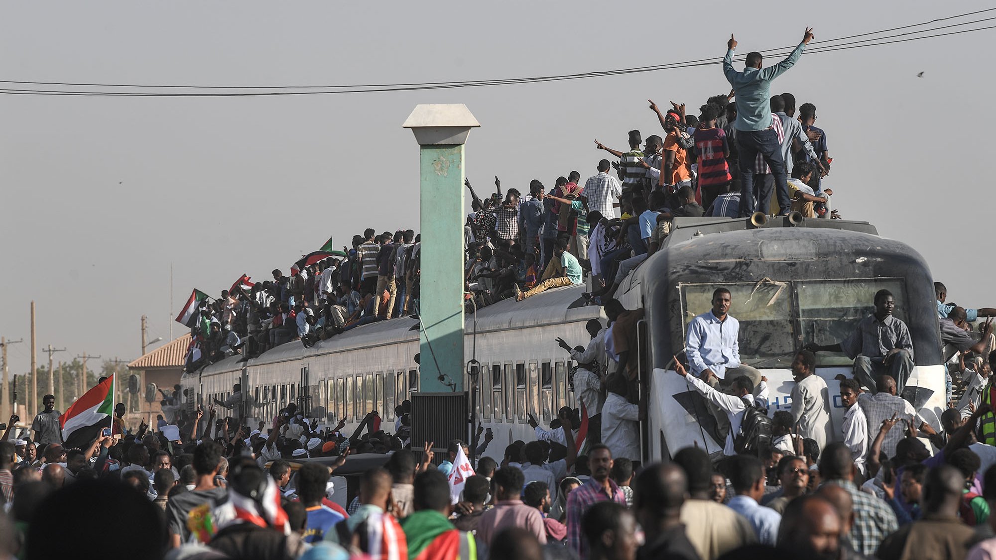 L'image montre une grande foule de personnes rassemblées autour et sur un train. Beaucoup de gens se tiennent sur le toit du train, tandis que d'autres s'accrochent aux côtés. L'atmosphère semble vibrante et énergique, probablement liée à une manifestation ou un événement important. En arrière-plan, on peut apercevoir des bâtiments et des infrastructures typiques d'une ville. La scène dégage un sentiment de solidarité et de détermination parmi les participants.
