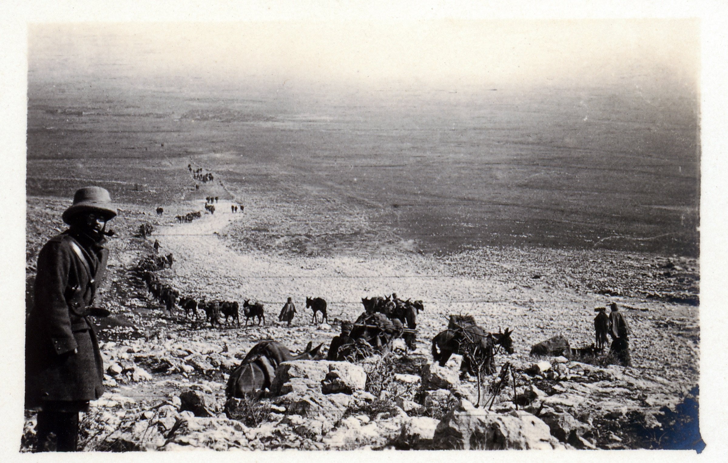 در این تصویر، مردی با کلاه و پوشاک سنتی در حال ایستادن در کنار یک شیب سنگی دیده می‌شود. در پس‌زمینه، گروهی از انسان‌ها و حیوانات (احتمالاً خرها یا اسب‌ها) در حال حرکت در یک مسیر سنگی به سمت پایین هستند. مناظر اطراف خشک و کوهستانی به نظر می‌رسد و تصویر حالتی قدیمی و سیاه و سفید دارد که به آن احساس تاریخ را القا می‌کند. فضای تصویر و نوع پوشش افراد نشان‌دهنده یک فعالیت یا سفر است.