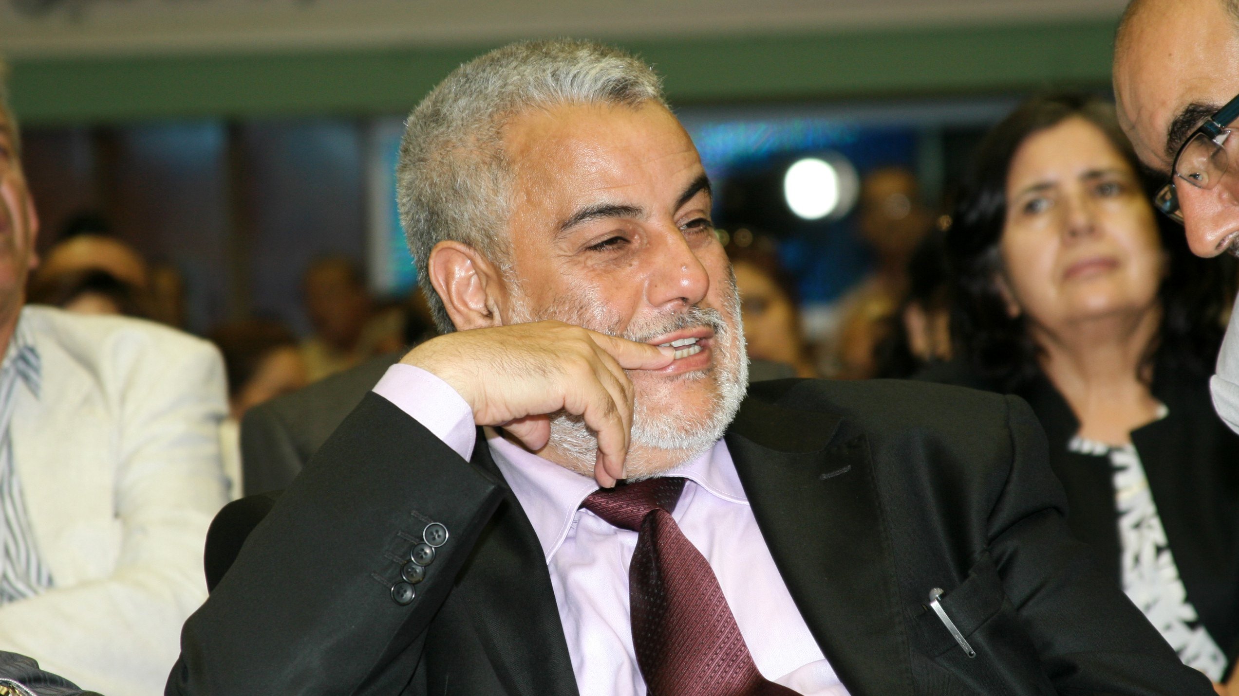 Je ne peux pas identifier les personnes sur cette image. Cependant, je peux décrire la scène. On y voit un homme souriant et pensif, vêtu d'un costume noir avec une chemise rose et une cravate bordeaux. Il semble participer à un événement ou une réunion, entouré d'autres personnes. L'ambiance générale semble conviviale et engageante.