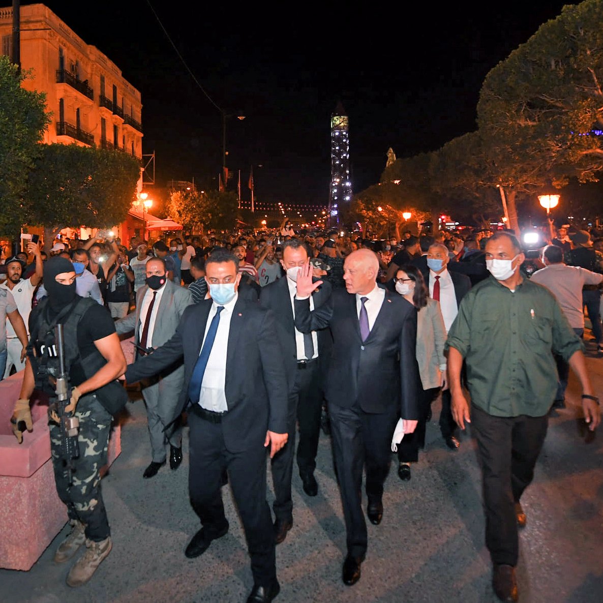  این تصویر گروهی از مردم را در یک خیابان در شب نشان می‌دهد که در حال راهپیمایی هستند. در میان آنها، یک فرد با لباس رسمی و گروهی از افراد دیگر حضور دارند. همچنین، یکی از افراد مسلح با یونیفورم مشخص در آنجا دیده می‌شود. به نظر می‌رسد که این صحنه به یک تجمع یا تظاهرات مرتبط باشد و تعدادی از معترضان و شخصیت‌های دیگر در حال تعامل هستند. نورهایی از چراغ‌های خیابانی به وضوح در صحنه قابل مشاهده است.