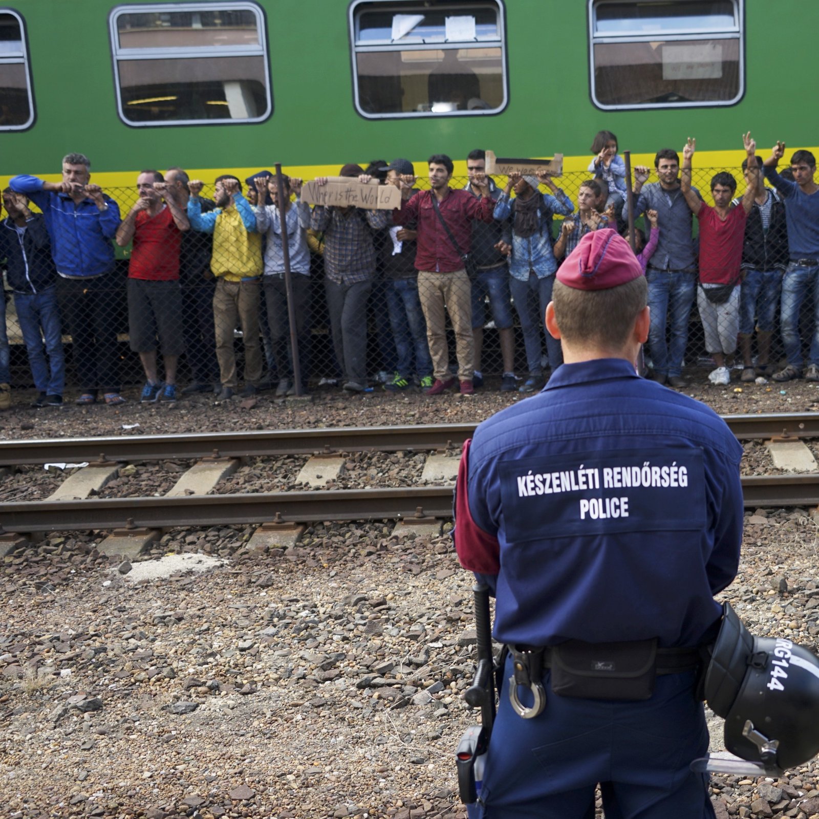 The image shows a scene at a train station where a police officer is facing a group of individuals. The officer is dressed in a uniform with the word "POLICE" clearly visible on the back. In front of the officer, there is a crowd of people, some holding signs, who appear to be expressing their discontent or making a statement. The background features a green train, and there are railway tracks visible, indicating that this is a transportation hub. The atmosphere seems tense, with the officer standing guard as the crowd raises their hands, possibly in protest or to seek attention.
