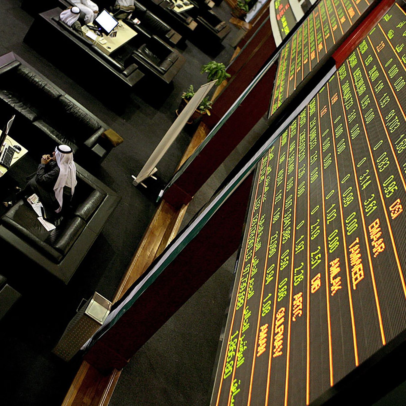 La imagen muestra una sala de operaciones en un mercado financiero. Hay pantallas grandes que exhiben datos sobre acciones y precios en un formato digital. En el fondo, se pueden ver personas sentadas en cómodos sofás, algunas de las cuales parecen estar revisando información en computadoras. El ambiente es moderno y está diseñado para brindar comodidad a los traders. La iluminación es suave y se puede notar un ambiente de concentración y actividad relacionada con el comercio de valores.