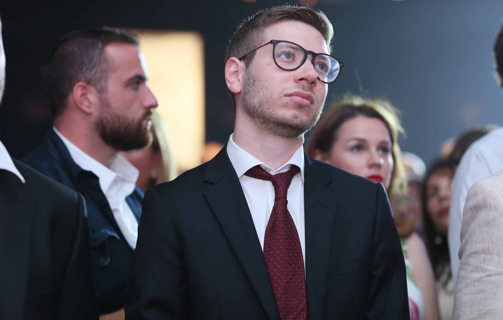 الصورة تُظهر مجموعة من الأشخاص في حدث اجتماعي أو عرض. في المقدمة، يبدو شاب يرتدي بذلة سوداء وقميصًا أبيض ورابطة عنق حمراء، وهو ينظر للأعلى بتعبير تأملي. خلفه يوجد العديد من الأشخاص، بعضهم يرتدي ملابس رسمية وآخرون بملابس أقل رسمية، مما يدل على تنوع الحضور. الإضاءة في الخلفية تعطي جوًا احتفاليًا.