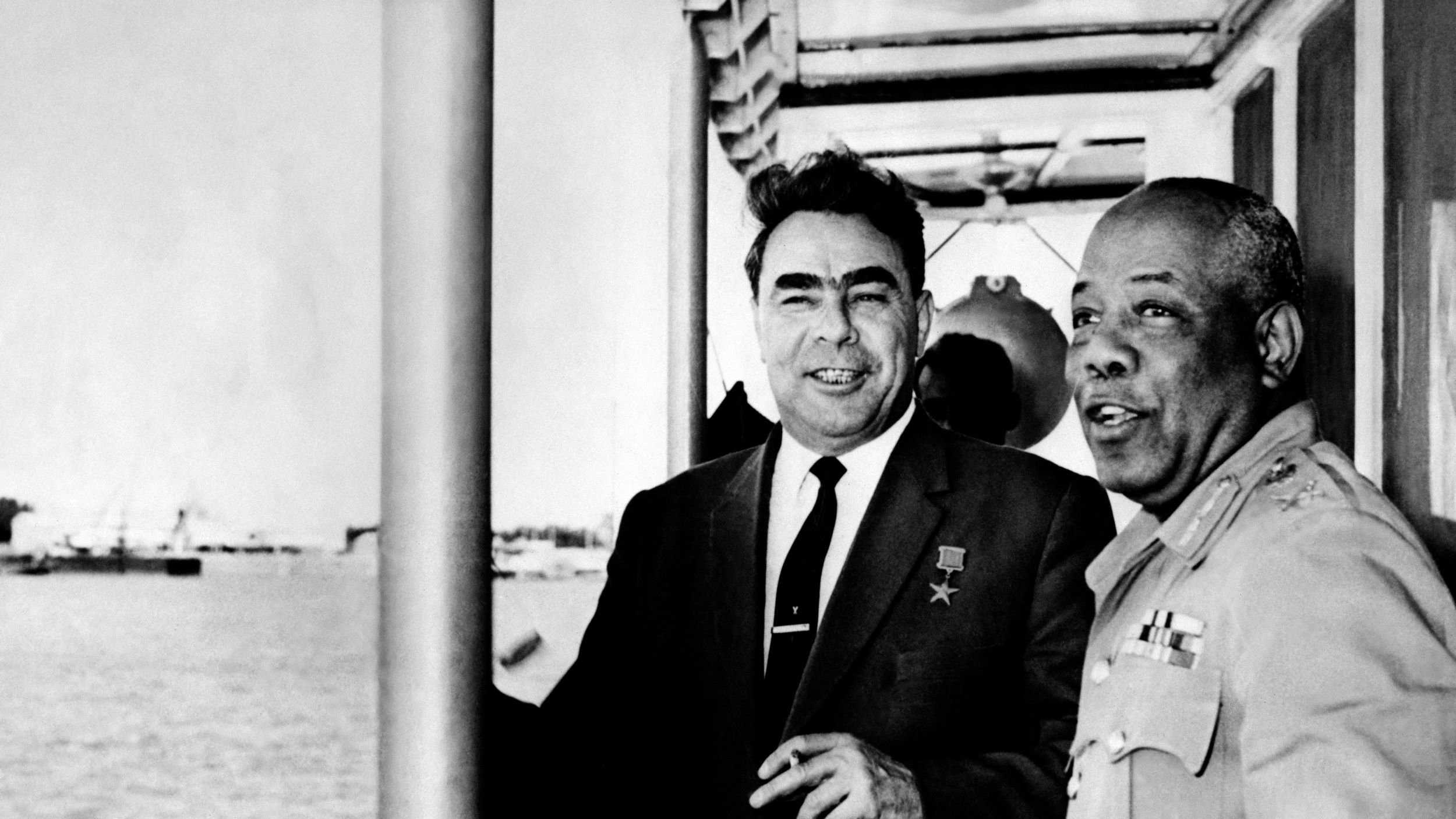 در تصویر، دو مرد در کنار یکدیگر ایستاده‌اند و به نظر می‌رسد که در نزدیکی آب هستند. یکی از آنها لباس رسمی و دکمه‌دار به تن دارد و بر سینه‌اش نشان یا مدالی دیده می‌شود. مرد دیگر نیز لباس نظامی بر تن دارد و در حال صحبت کردن یا تبادل نظر با مرد اول است. پس‌زمینه تصویر نمایی از آب و ممکن است قایق‌ها یا بنادر باشد. احساس کلی تصویر دوستانه و غیررسمی به نظر می‌رسد.