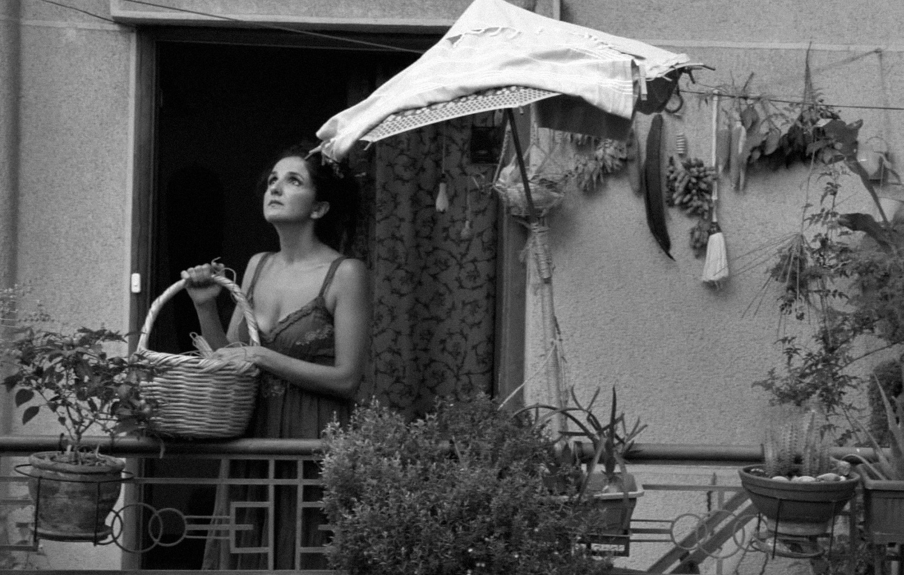 L'image montre une femme se tenant sur un balcon, tenant un panier dans ses bras. Elle semble contemplative, regardant au loin. Le balcon est décoré de plusieurs plantes et herbes suspendues. Un parasol en toile protège en partie la femme du soleil. L'image est en noir et blanc, ce qui lui donne une atmosphère nostalgique et mélancolique.